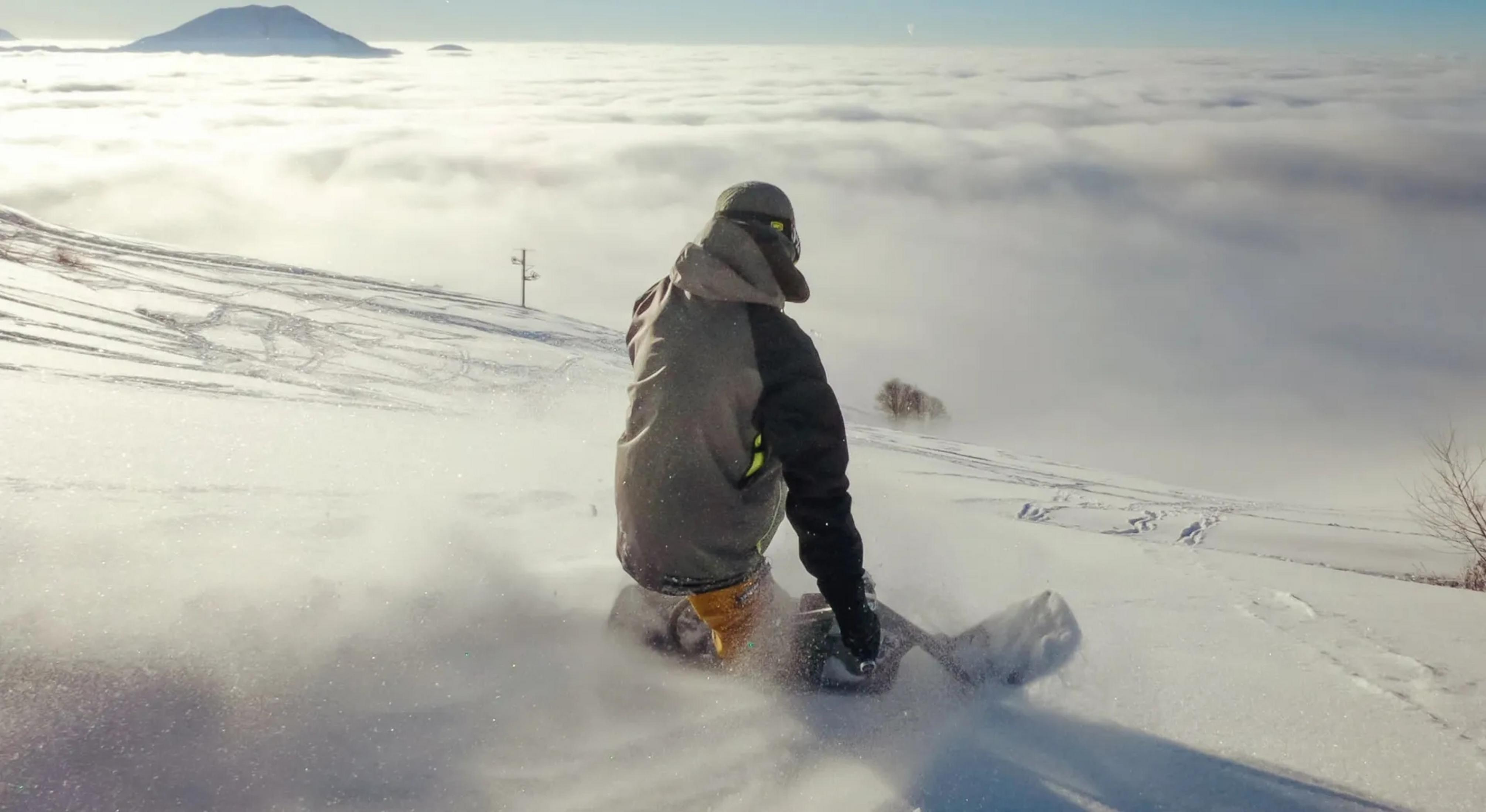 Snowboardzista jedzie przez puch z morzem chmur w tle.
