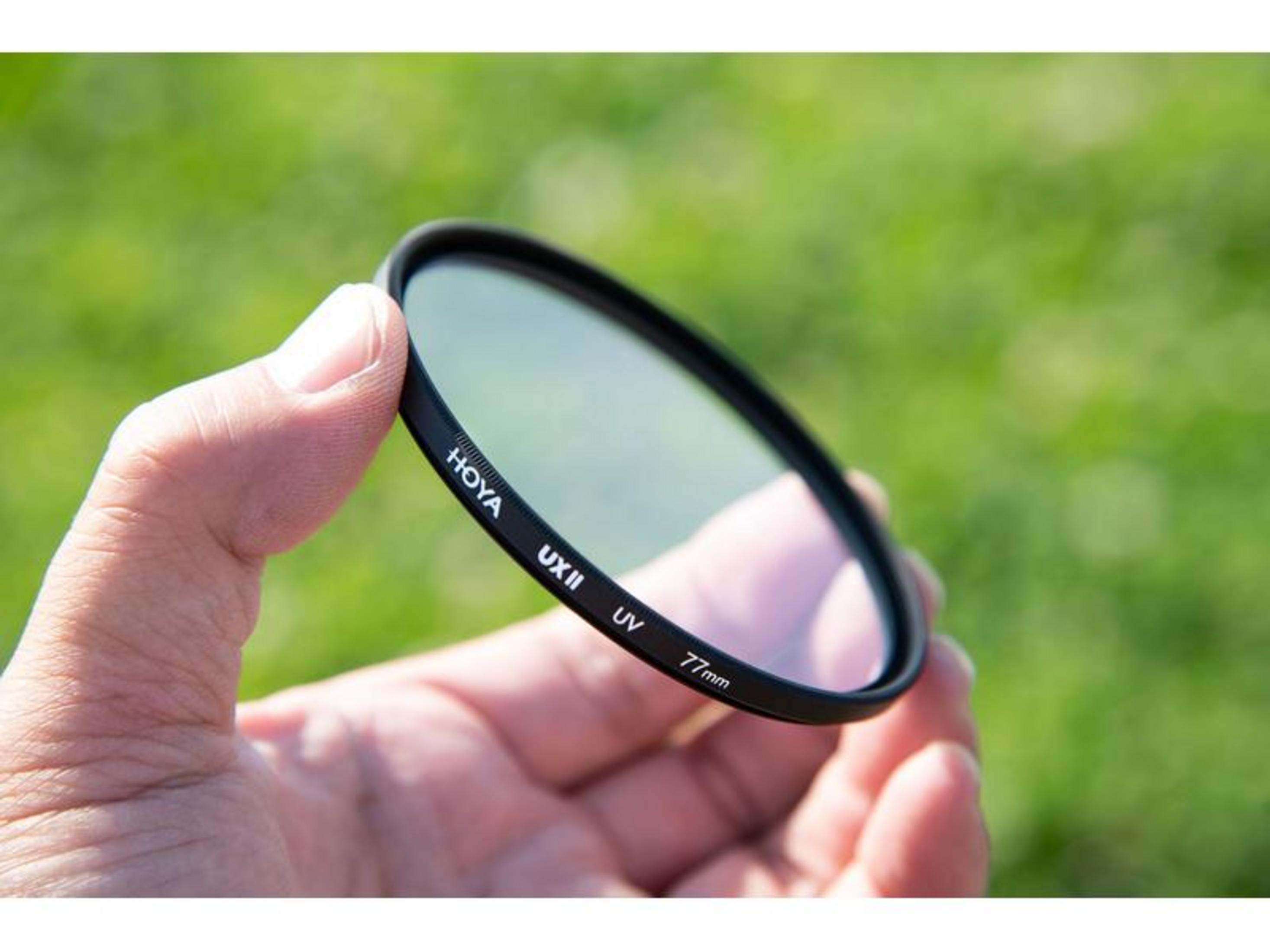 Eine Hand hält einen Hoya UX II UV 77mm Filter vor unscharfem grünen Hintergrund.