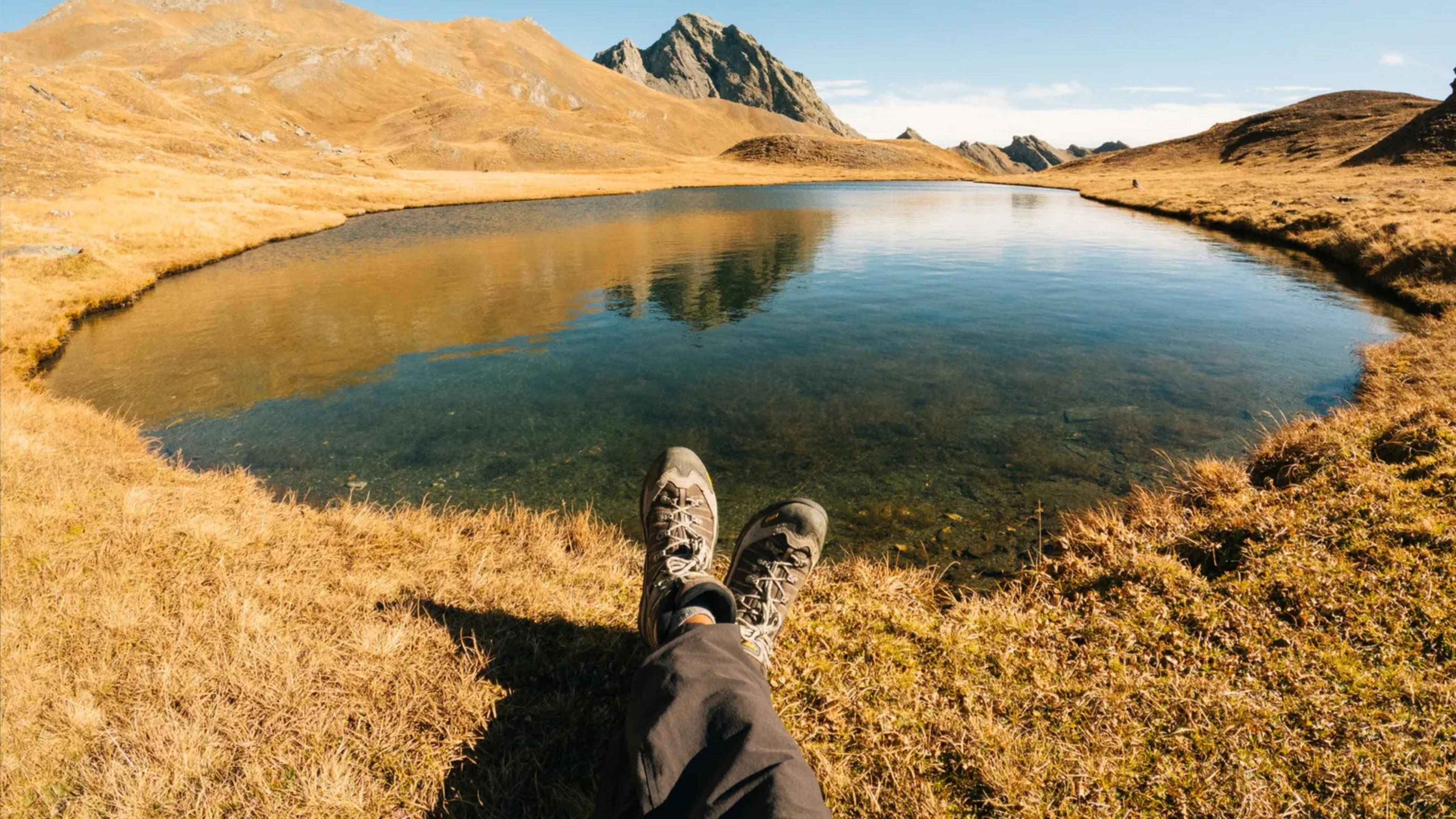 Nogi osoby w butach trekkingowych spoczywają na suchej trawie przy spokojnym górskim jeziorze.