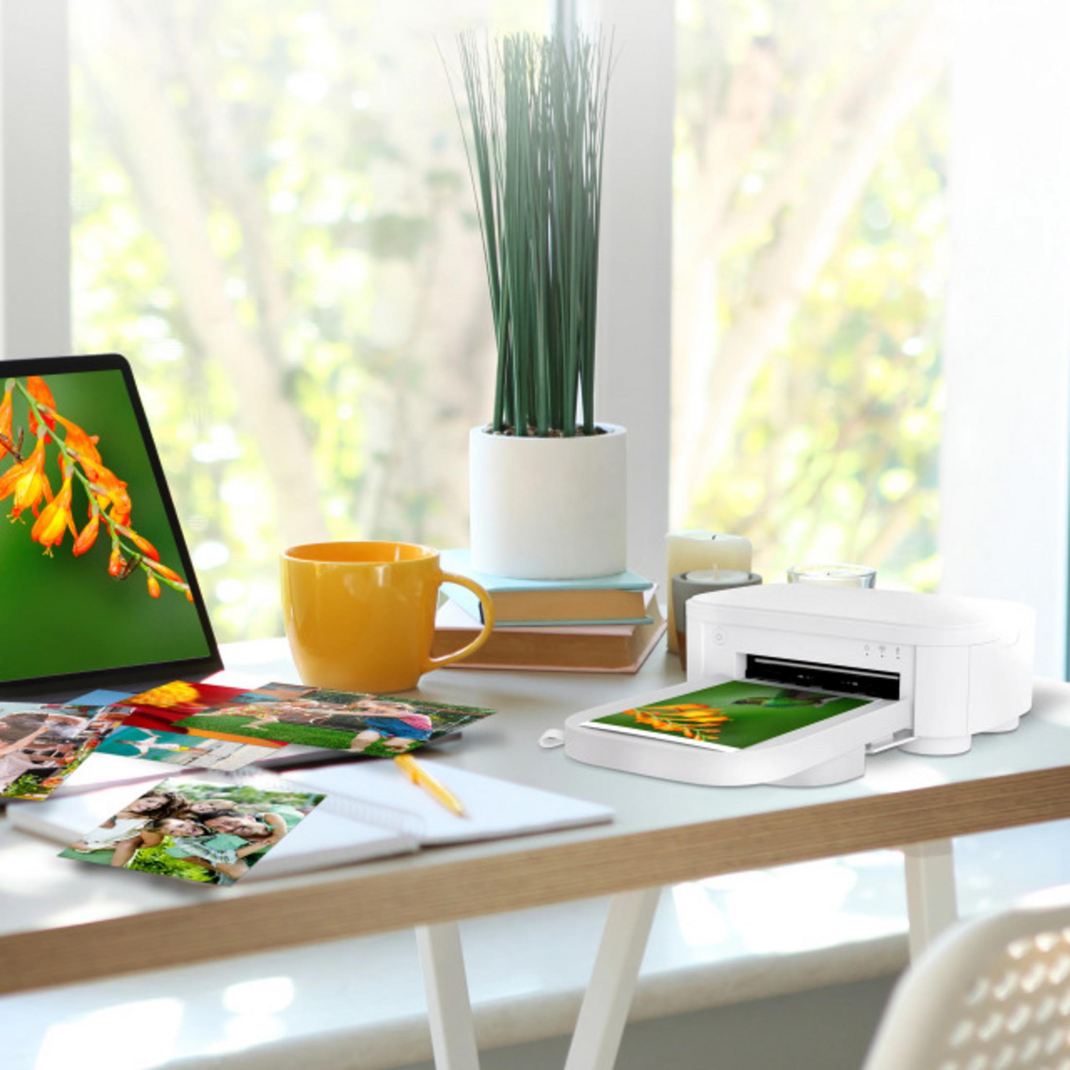 Laptop, żółty kubek, zdjęcia i drukarka na biurku.