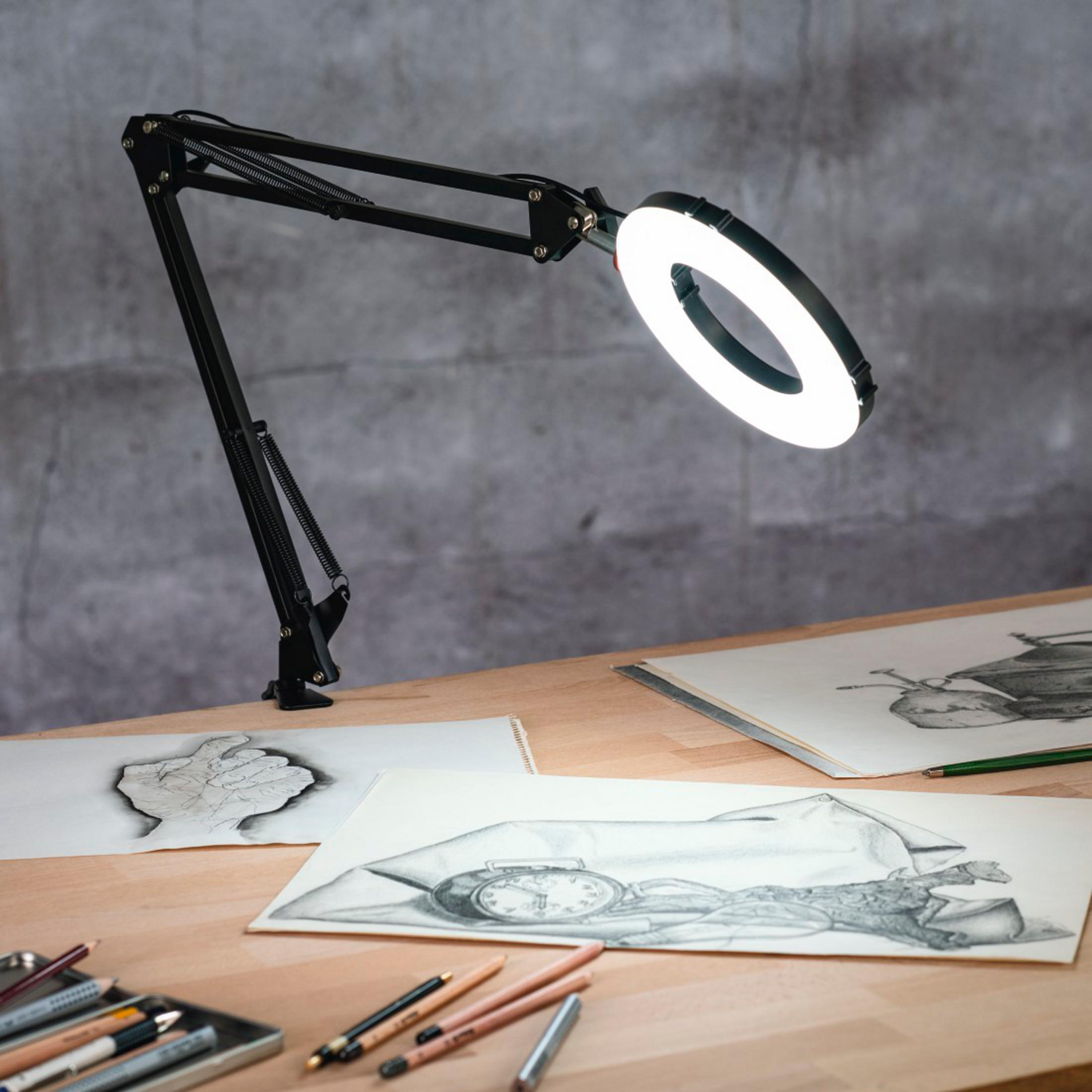 Ein schwarzer Schreibtischarm mit Ringlicht leuchtet auf einem Holztisch mit Skizzen.