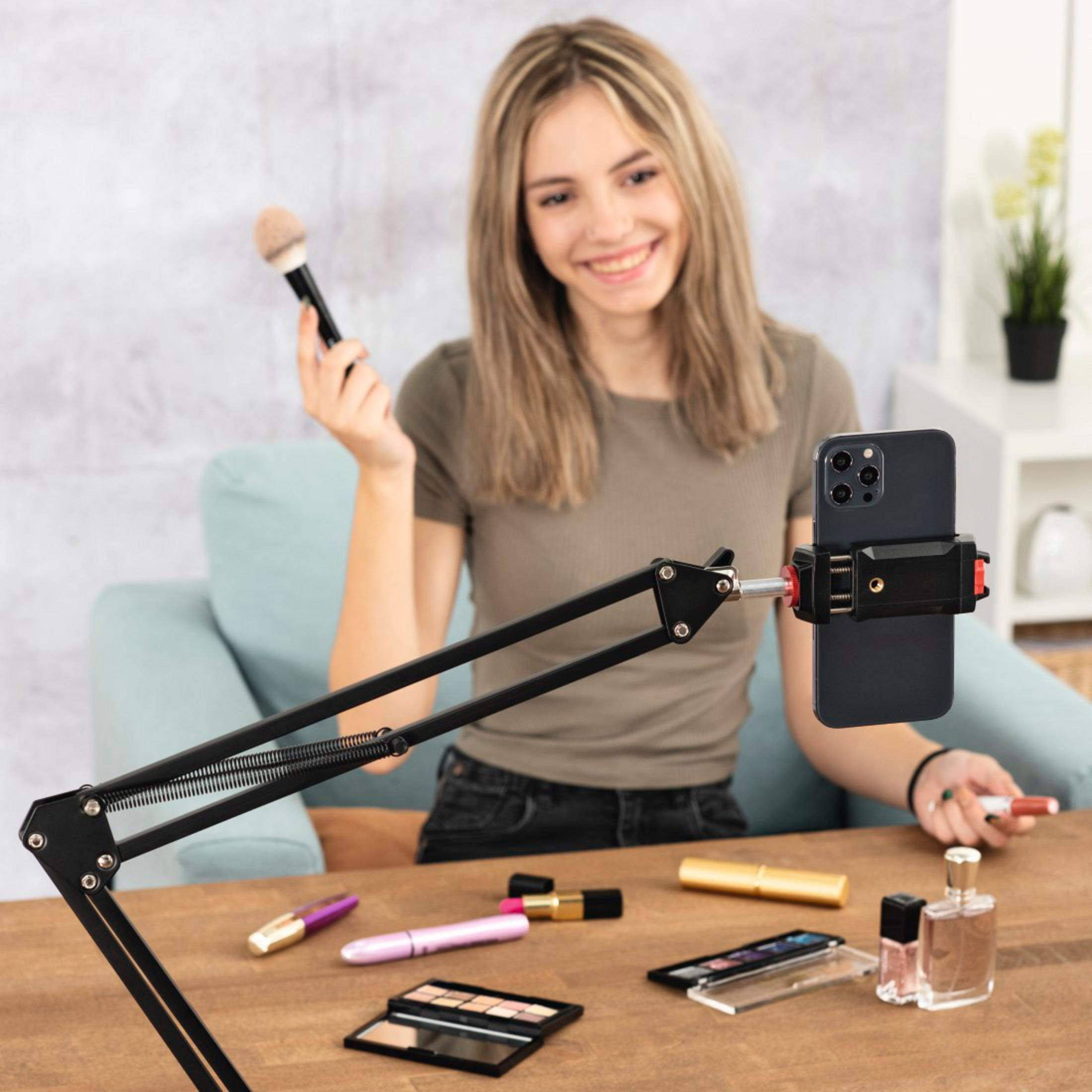 Eine Frau hält einen Pinsel und Lippenstift vor einem Handy, das auf einem Ständer montiert ist.
