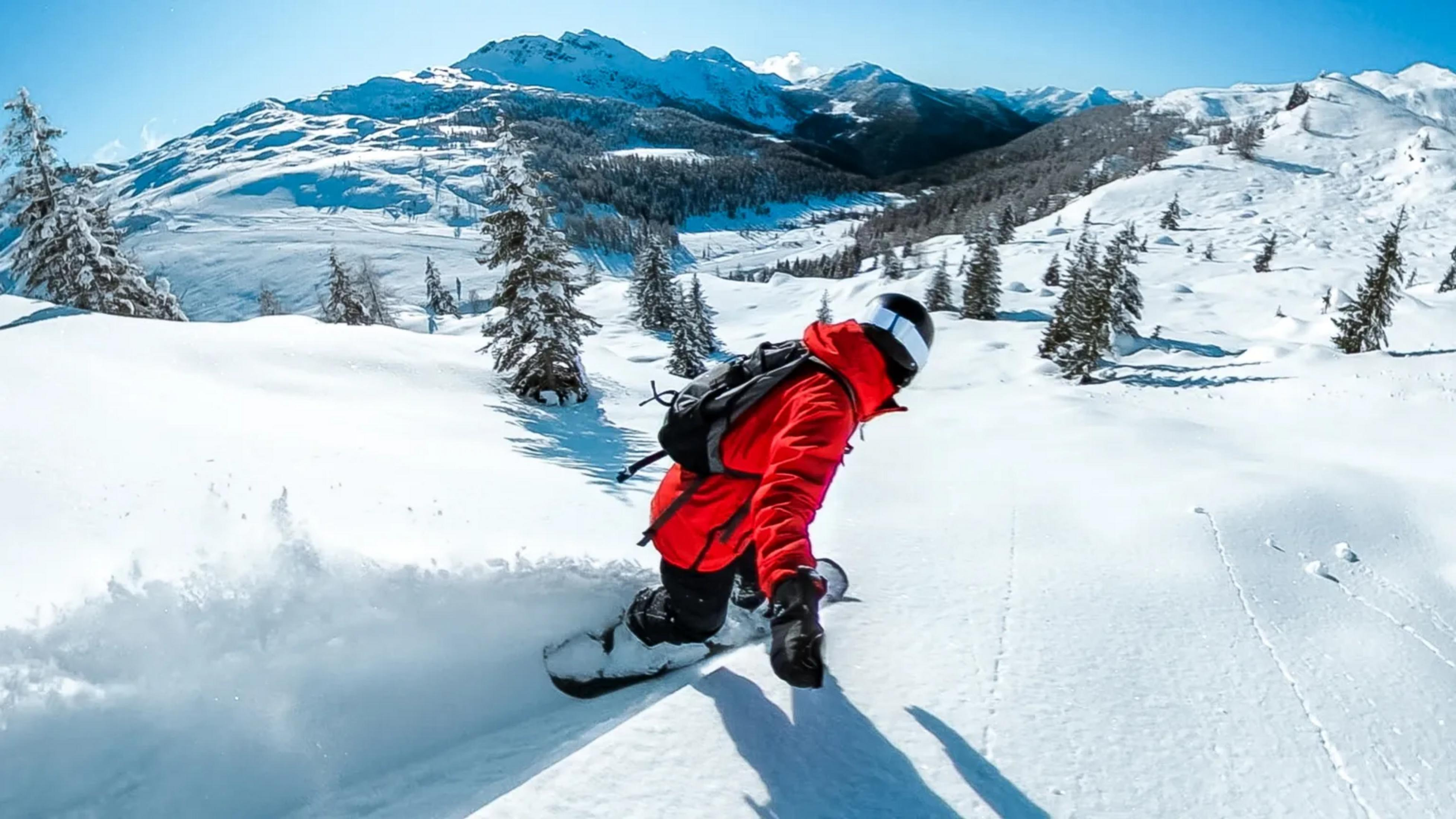 Snowboardzista w czerwonej kurtce jedzie po puchu, góry w tle.