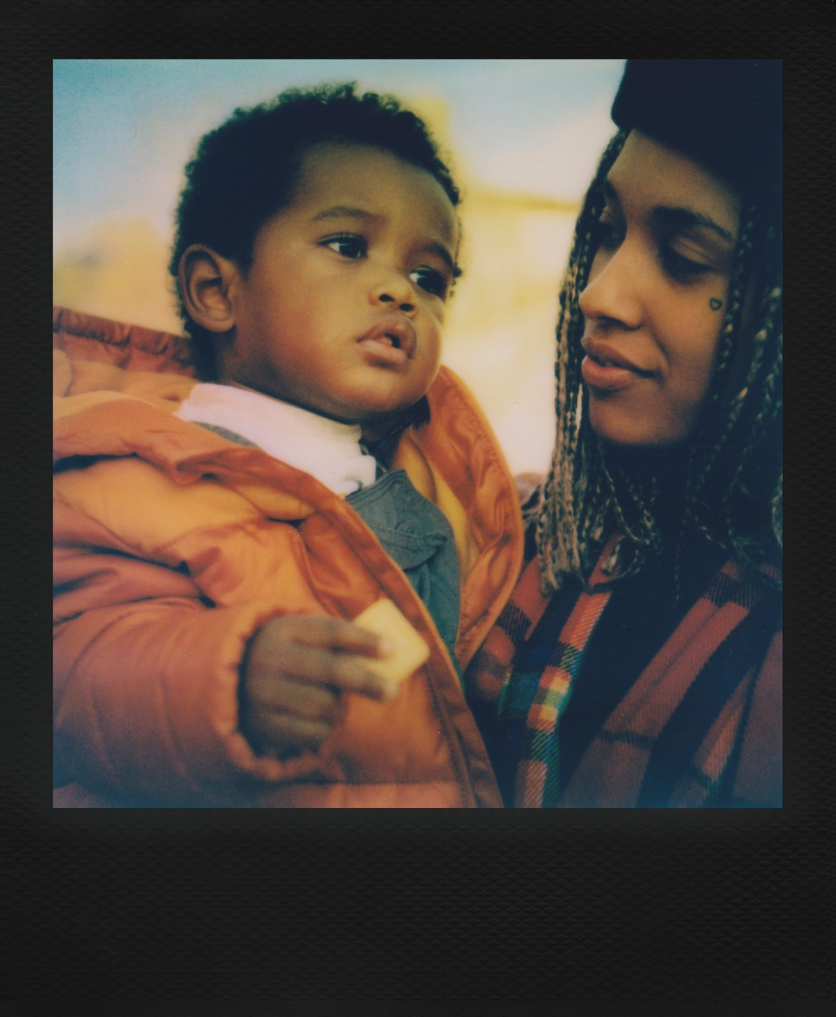 Maluch w pomarańczowej kurtce trzyma żółty przedmiot. Patrzy na niego osoba z warkoczami i w kratę.