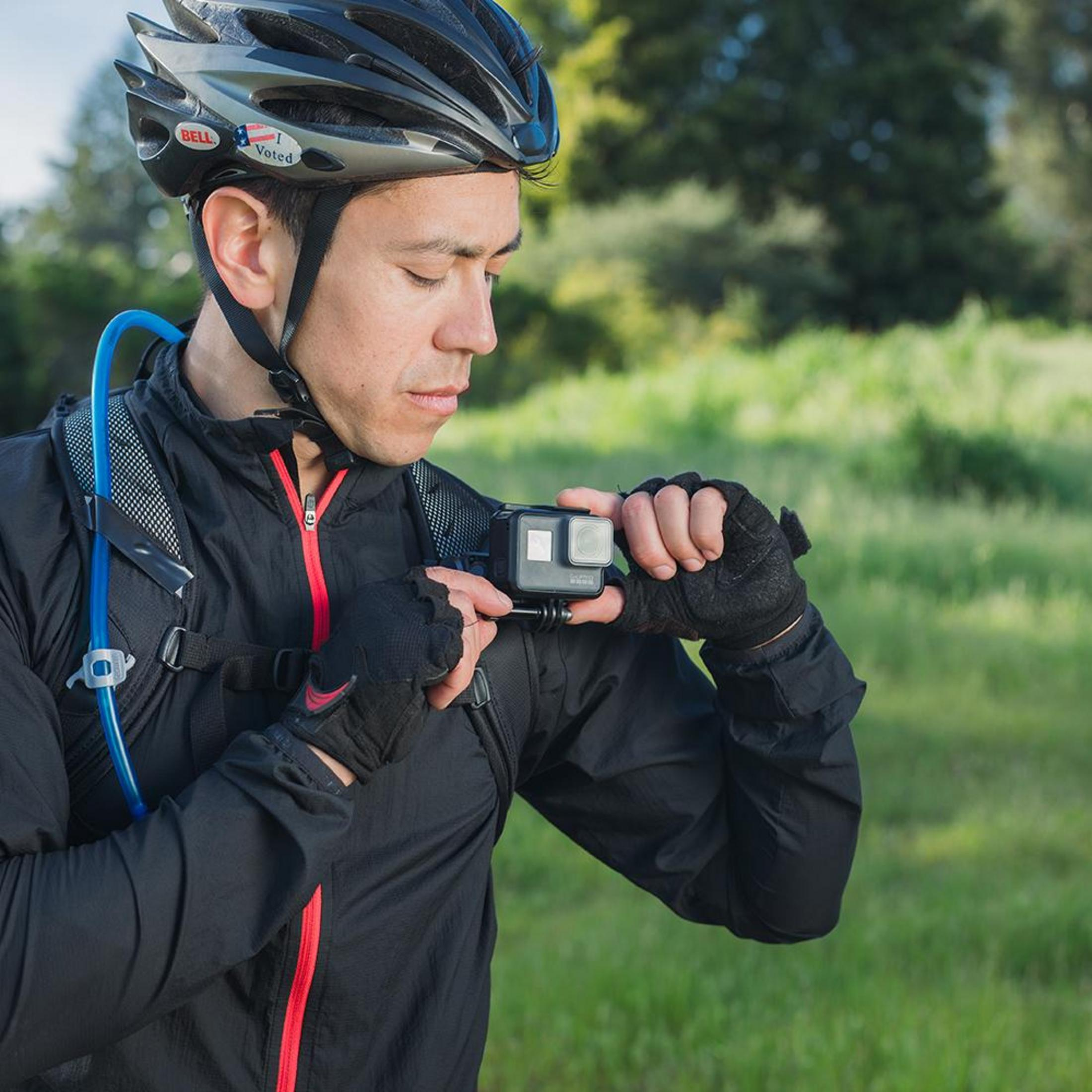 Un uomo con casco e guanti regola una fotocamera, con erba verde sullo sfondo.