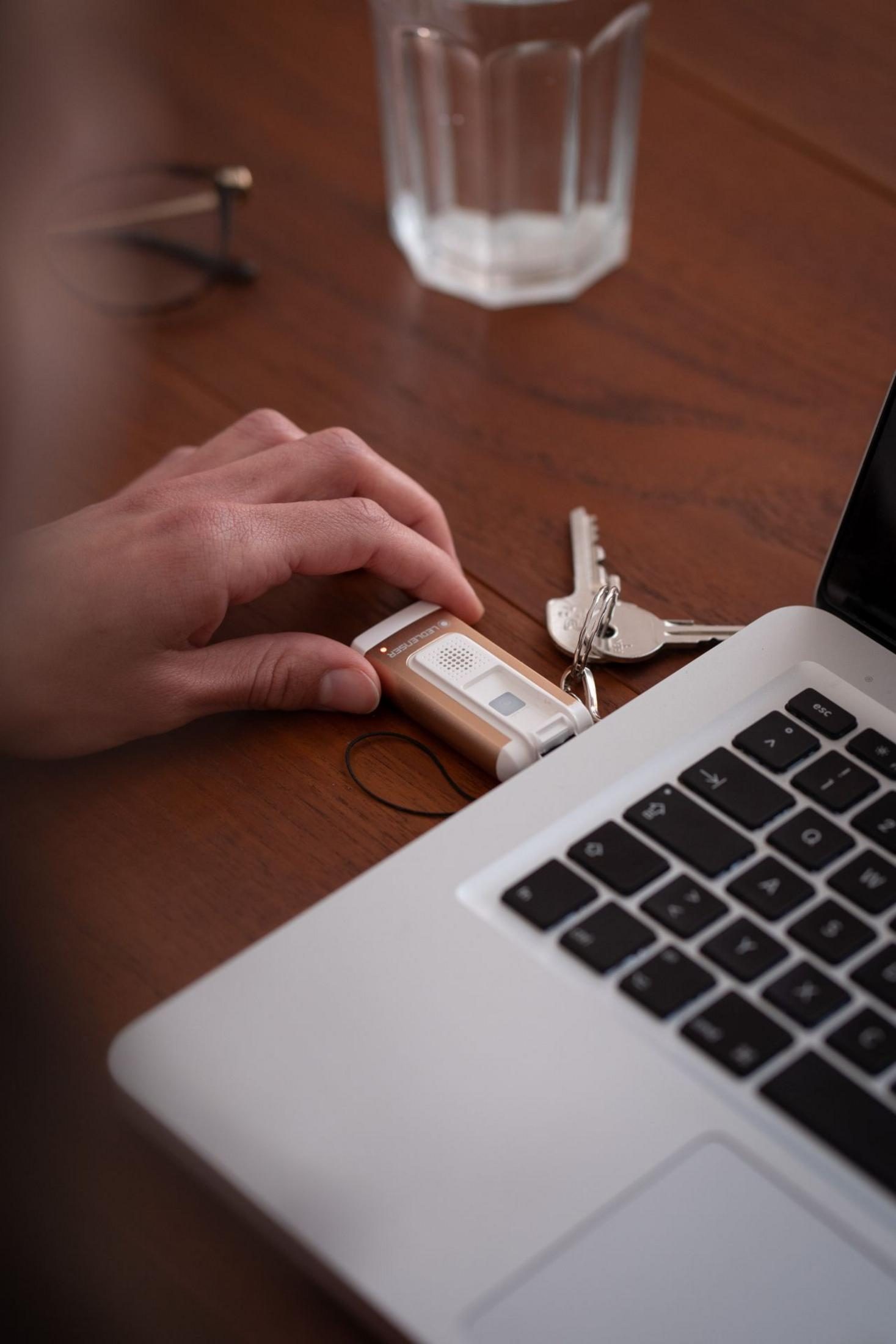 Dłoń sięga po małe, złote urządzenie obok laptopa, kluczy i szklanki.