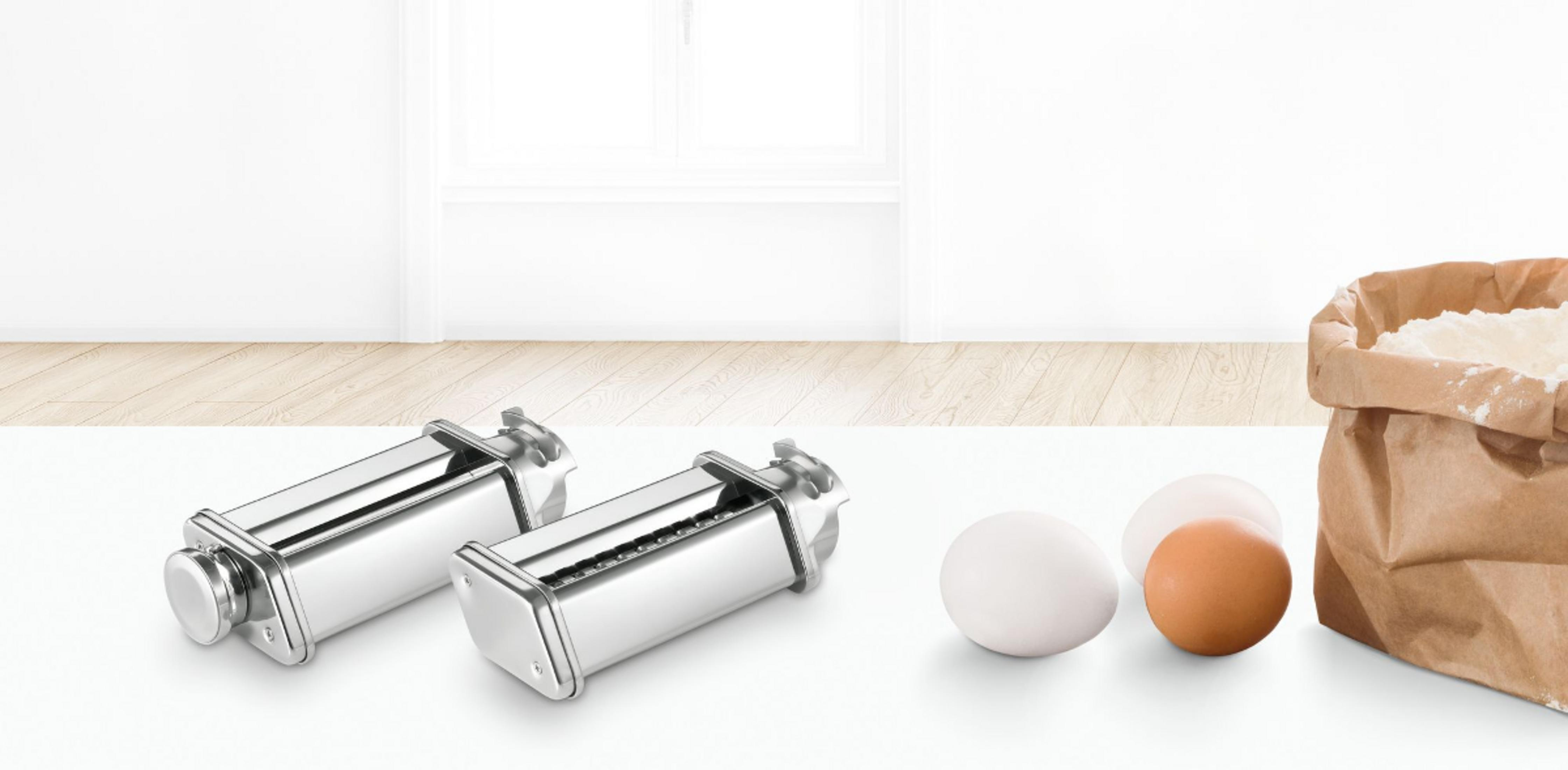 Dos máquinas de pasta, tres huevos y un saco de harina en mesa blanca.