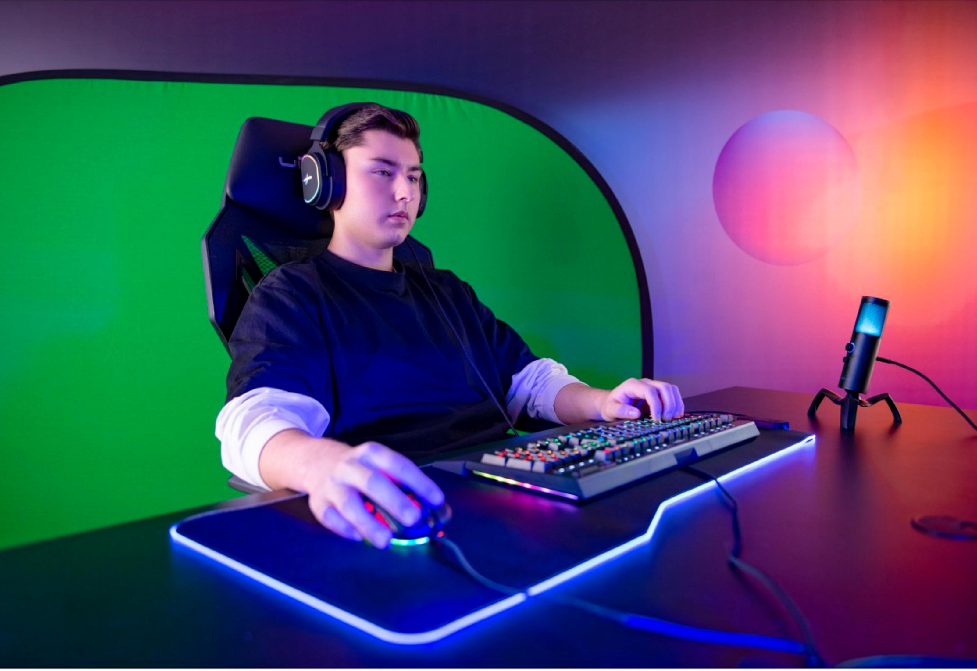 Ein Mann mit Kopfhörern spielt am PC in einem Raum mit grüner Leinwand.