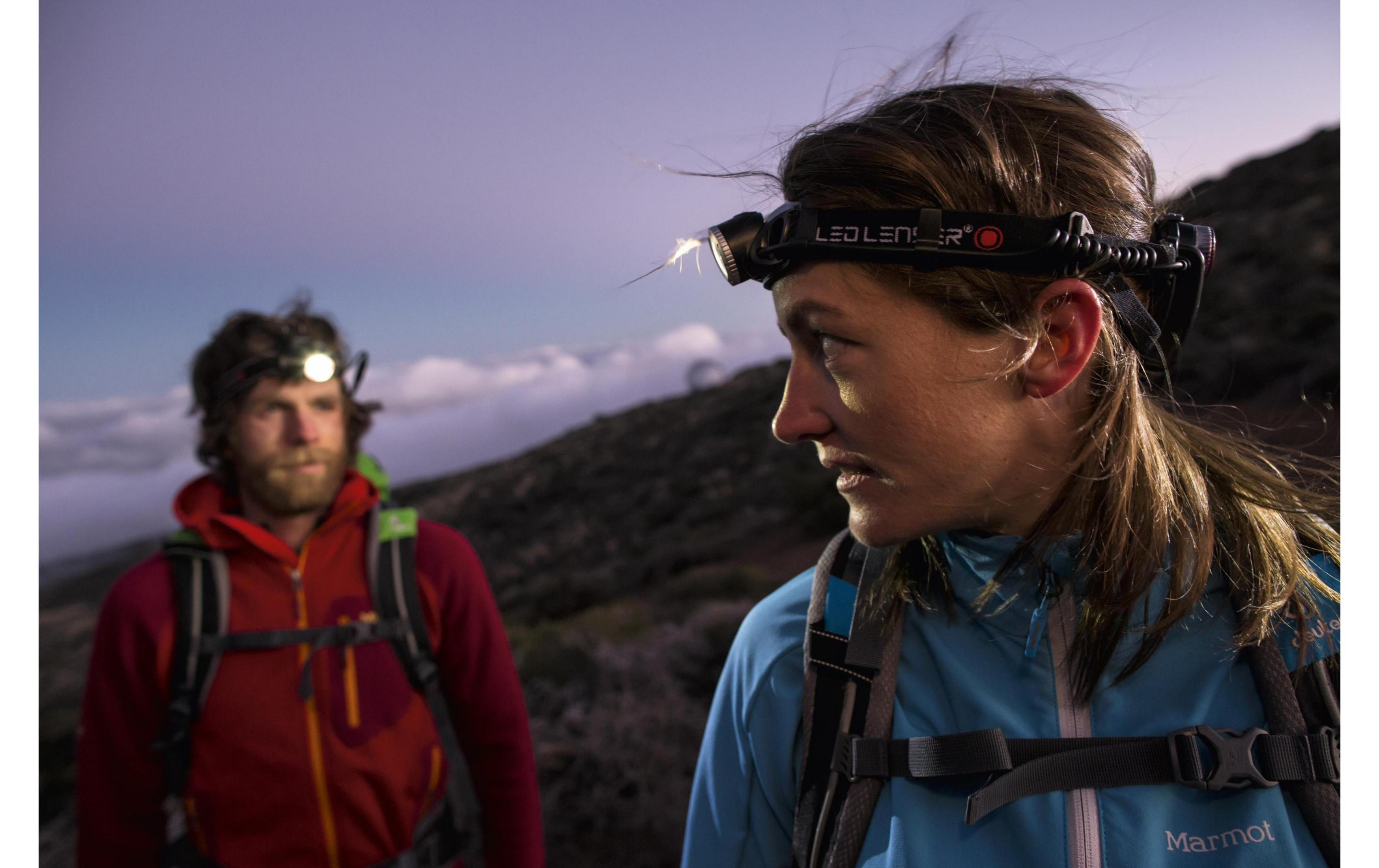 Deux randonneurs avec lampes frontales au crépuscule devant un paysage de montagne.