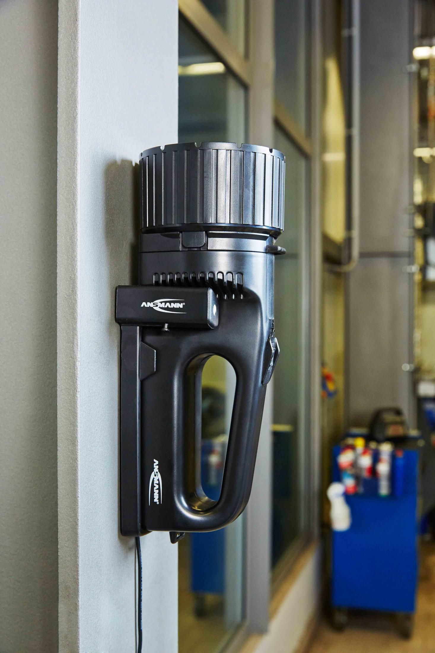 Schwarze Ansmann-Taschenlampe an Wand montiert, Werkzeugwagen im Hintergrund.
