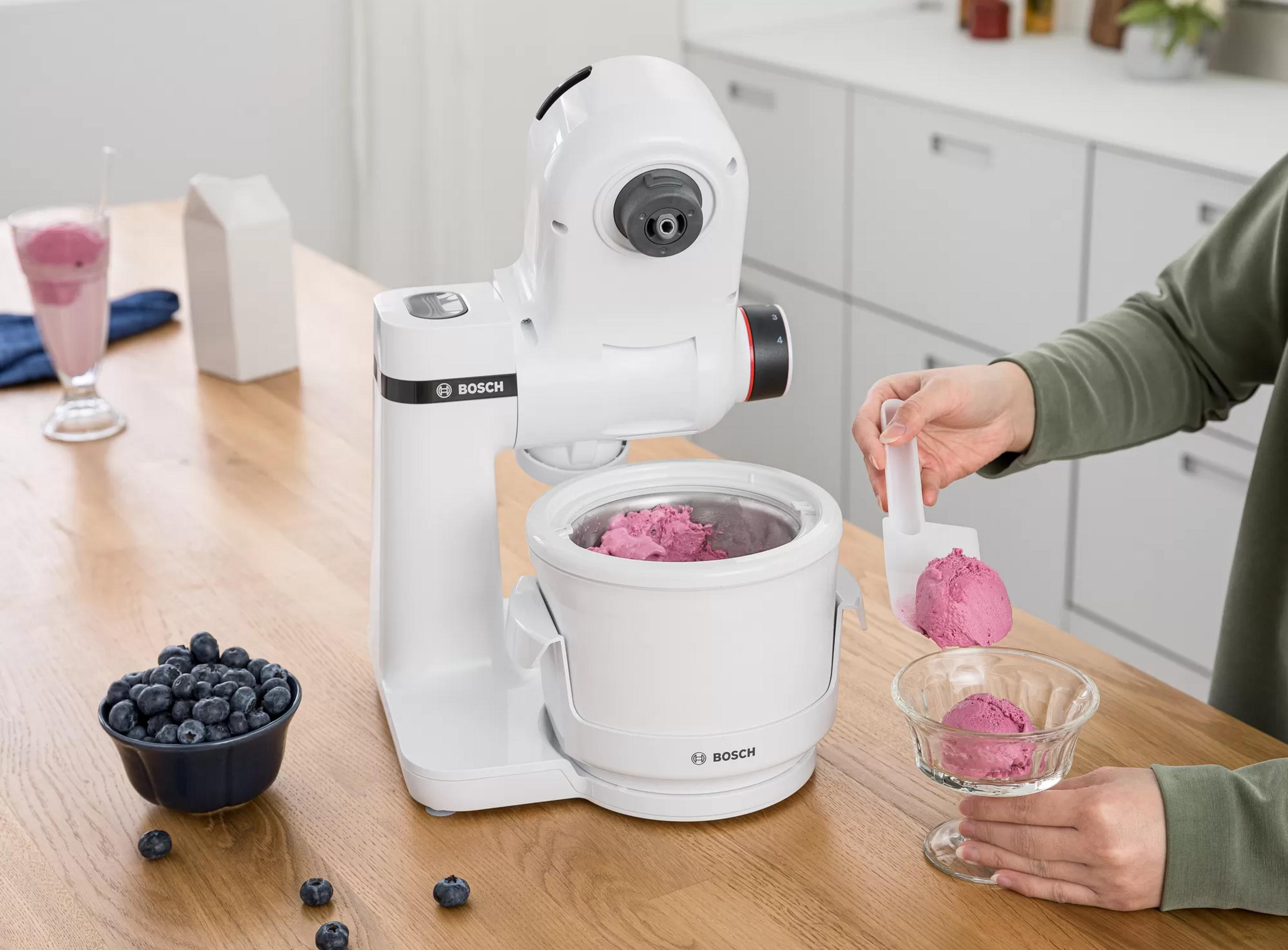 Eine weiße Eismaschine auf einem Holztisch macht Eis. Blaubeeren liegen daneben.
