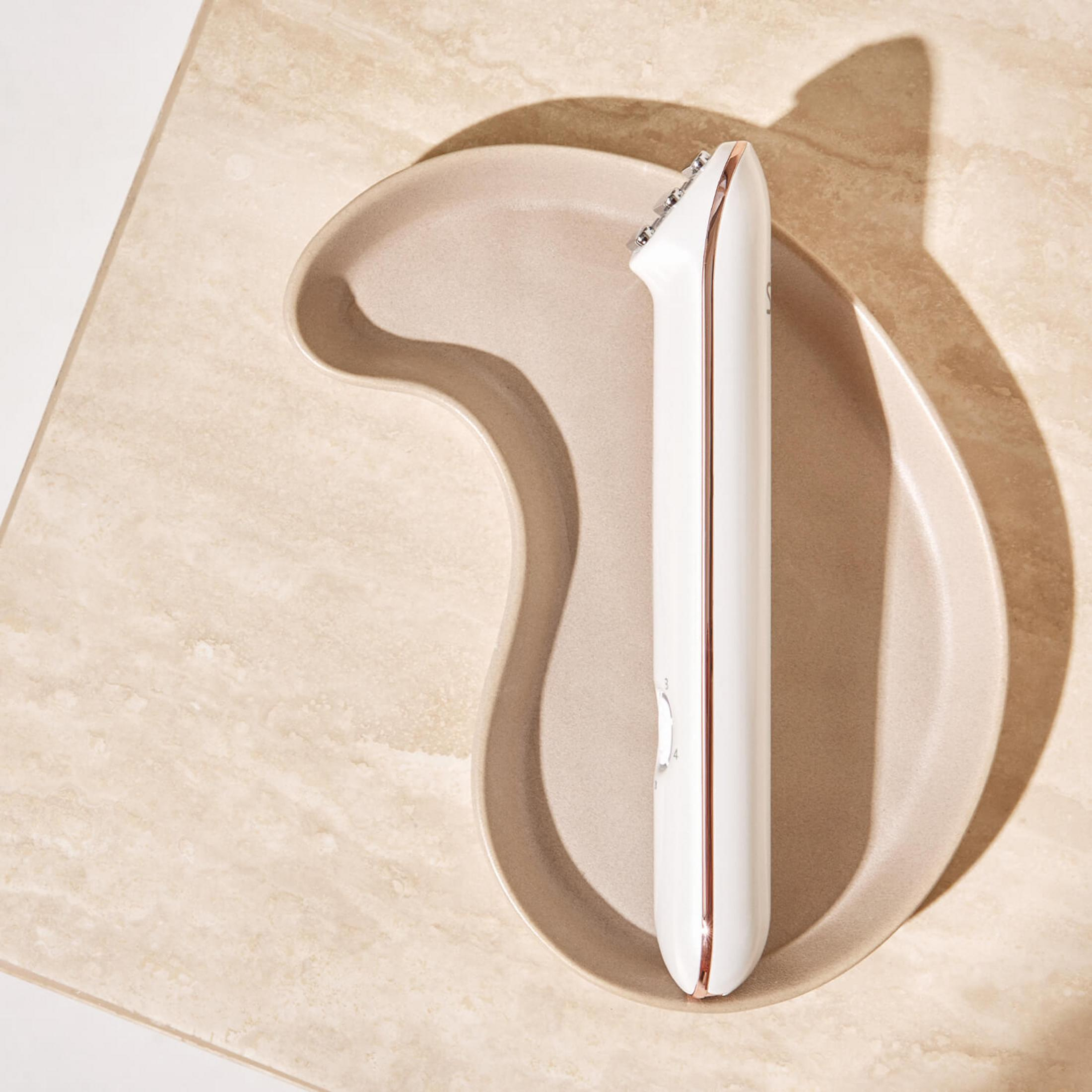 Ein weißer Beauty-Roller mit Roségold-Akzenten liegt auf einer geschwungenen, hellbraunen Schale.
