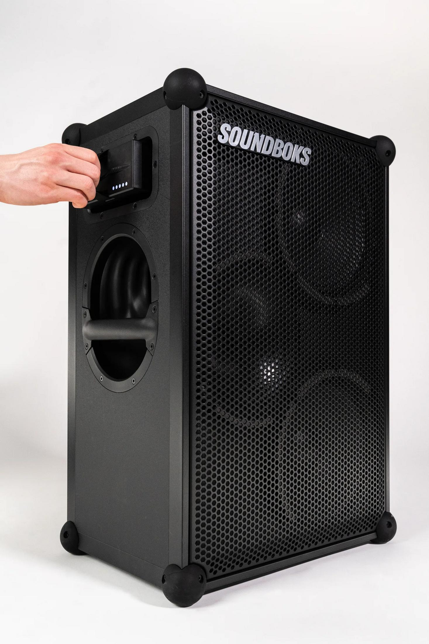 Schwarzer Lautsprecher mit Hand, die ein Kabel einsteckt. SOUNDBOKS Logo oben.