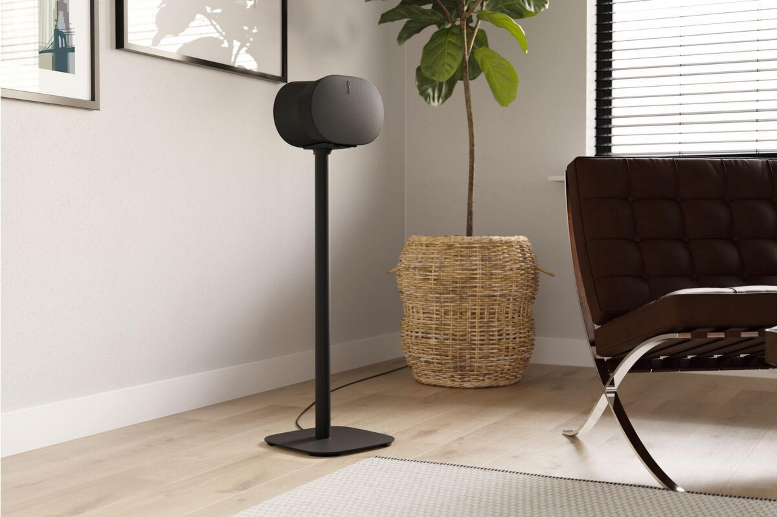 Czarny głośnik stoi na stojaku obok kosza i fotela.