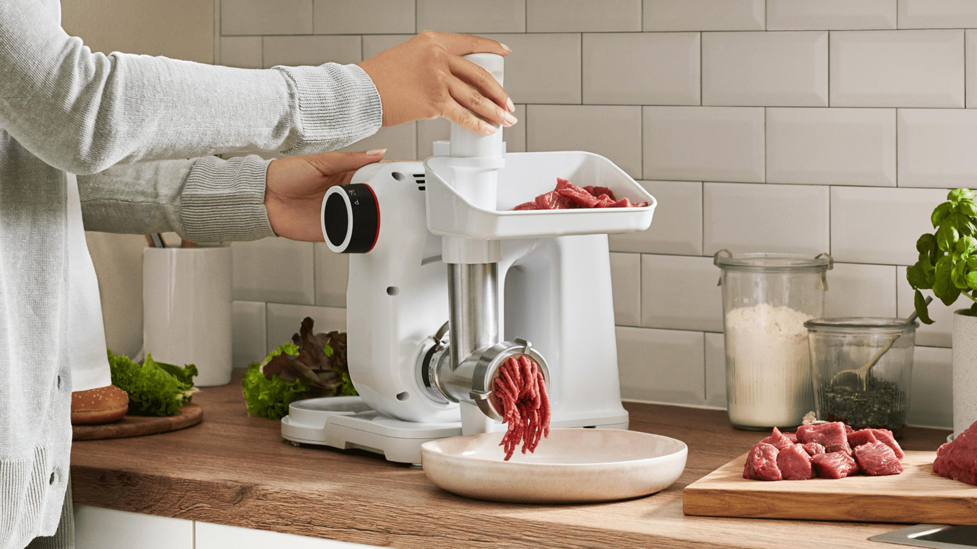 Eine Person benutzt einen Fleischwolf, um rohes Fleisch zu mahlen. Auf der Arbeitsplatte liegen weitere Zutaten.