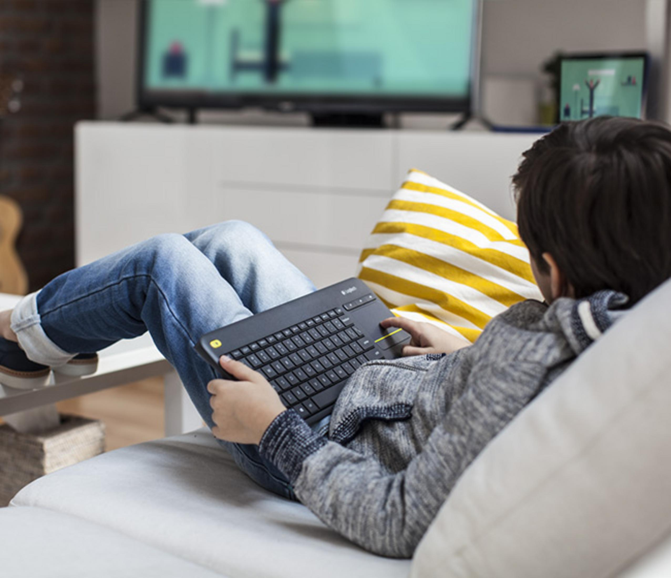 Chłopiec leży na sofie, używając klawiatury. W tle widać ekran telewizora.