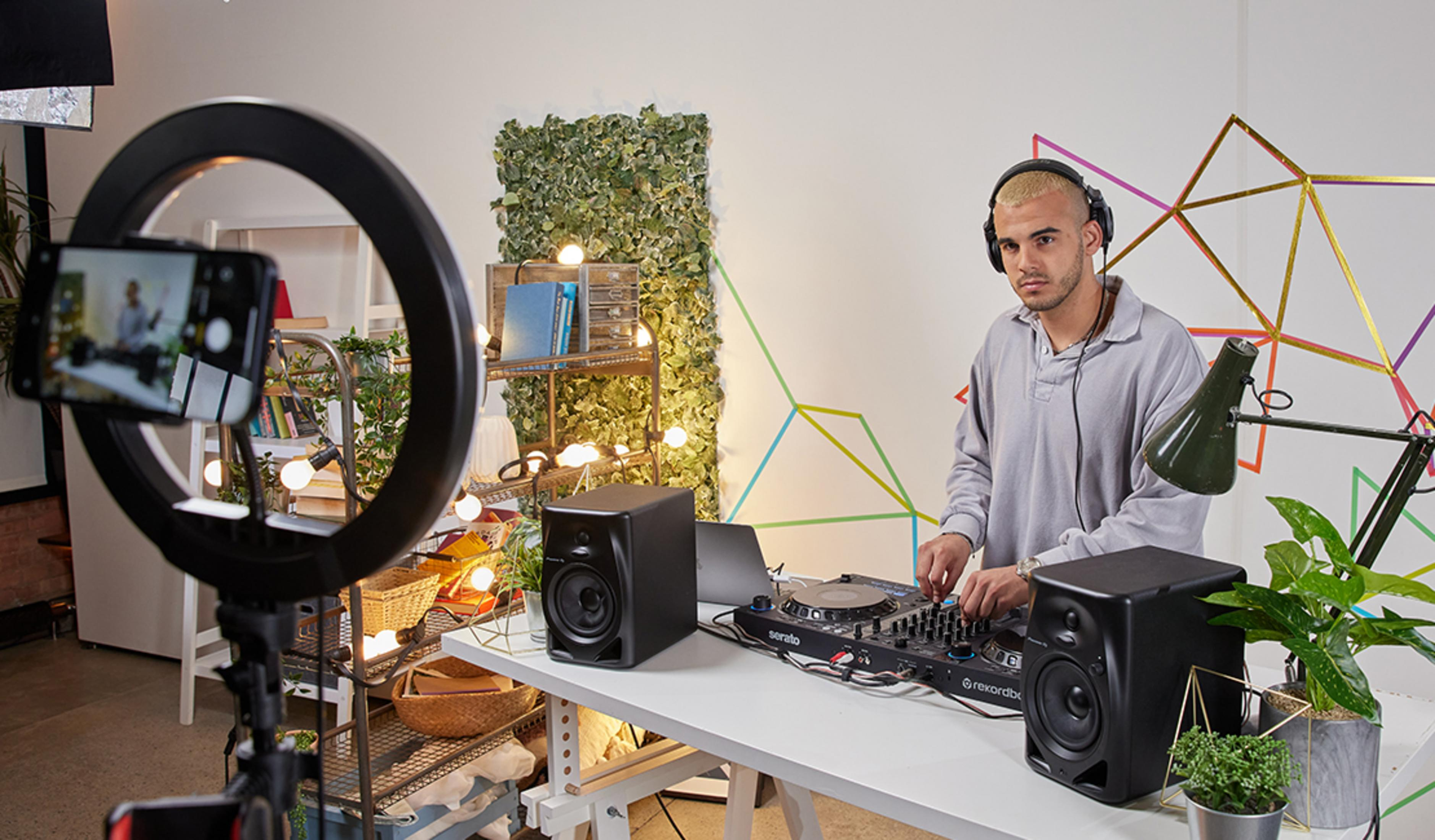A DJ wearing headphones plays music on a mixer, with a ring light camera in the foreground.