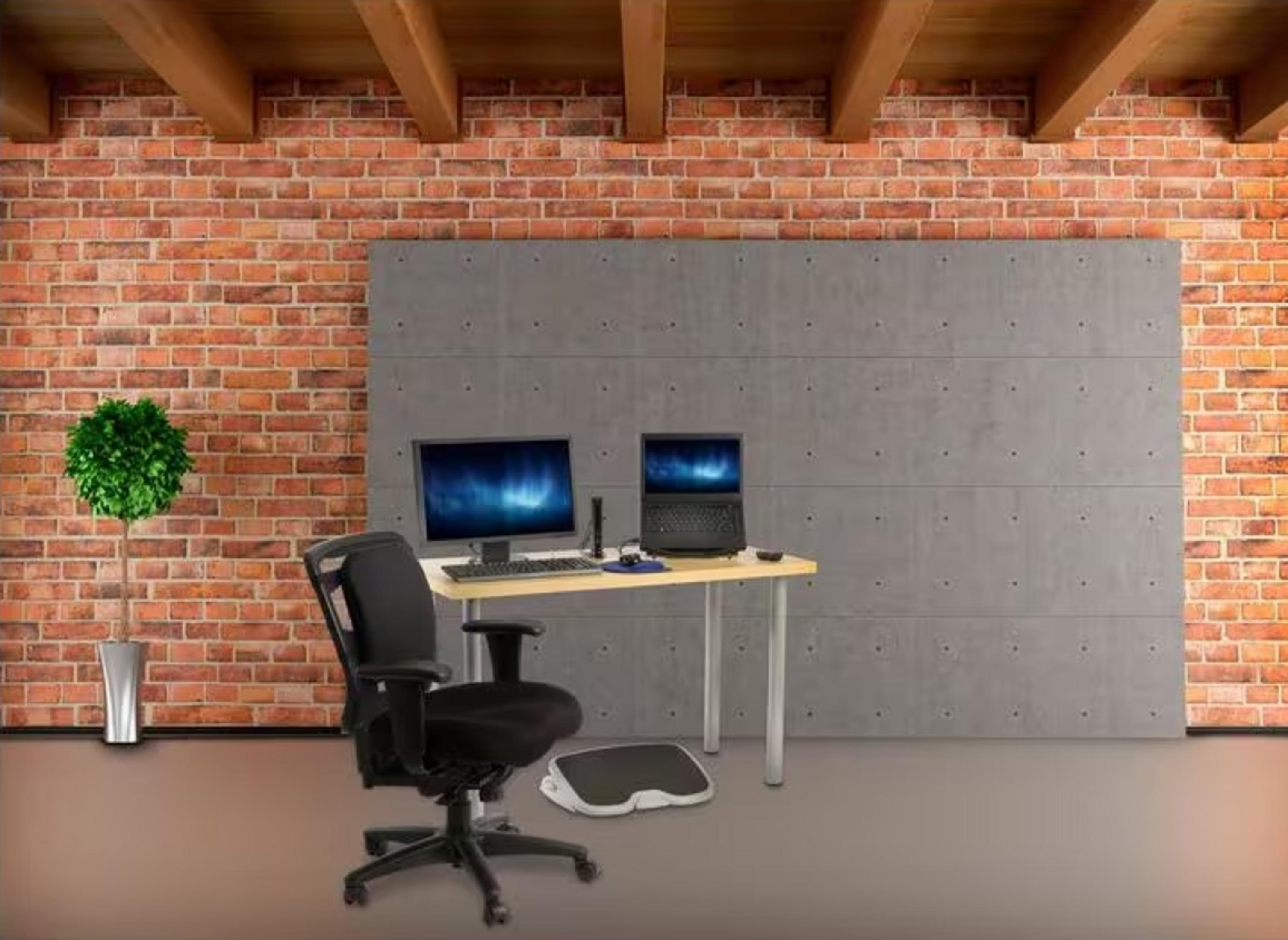 Büro mit Laptop, Monitor, Stuhl und Fußstütze vor Ziegel-/Betonwand.