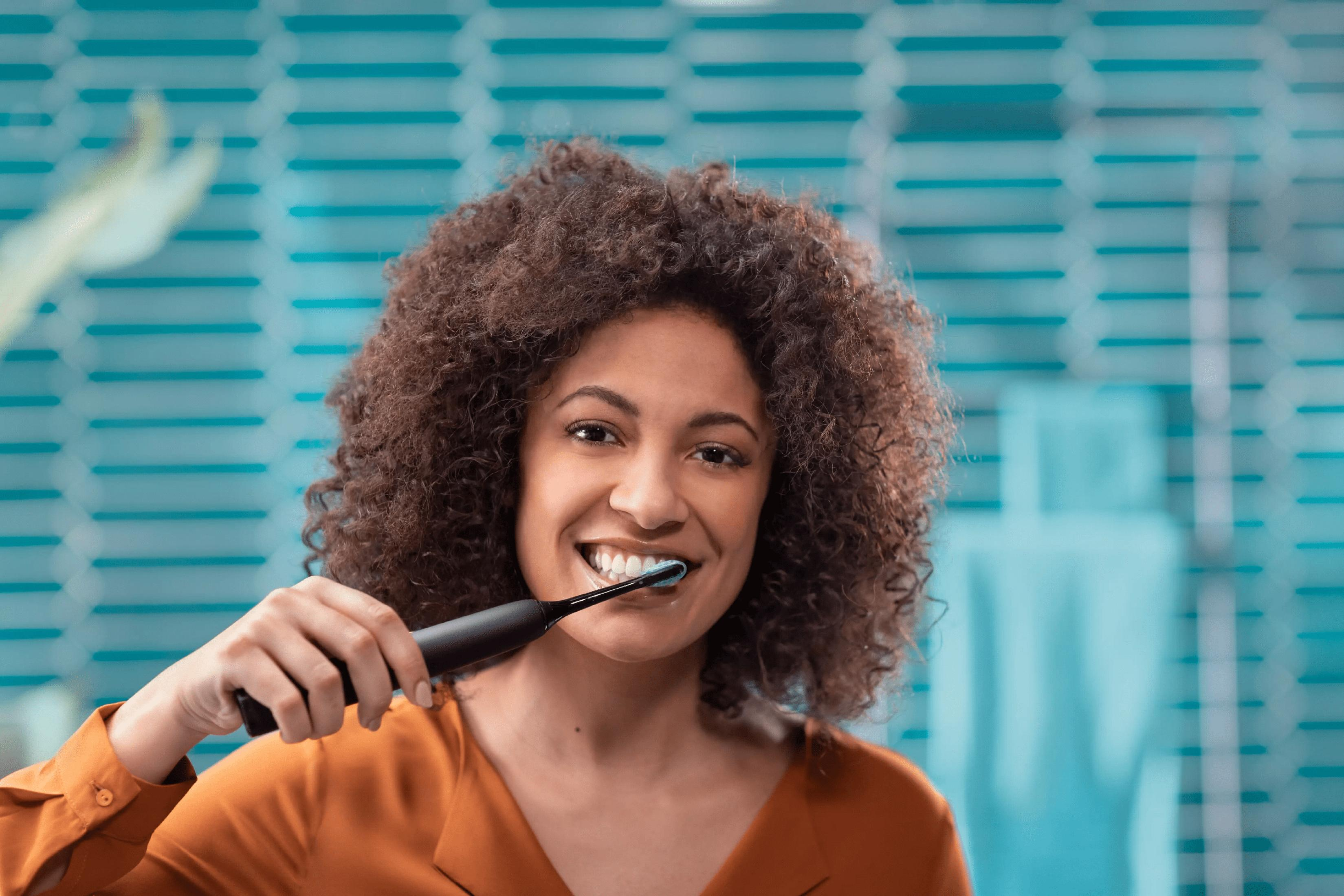 Kobieta z kręconymi włosami myje zęby czarną szczoteczką elektryczną.