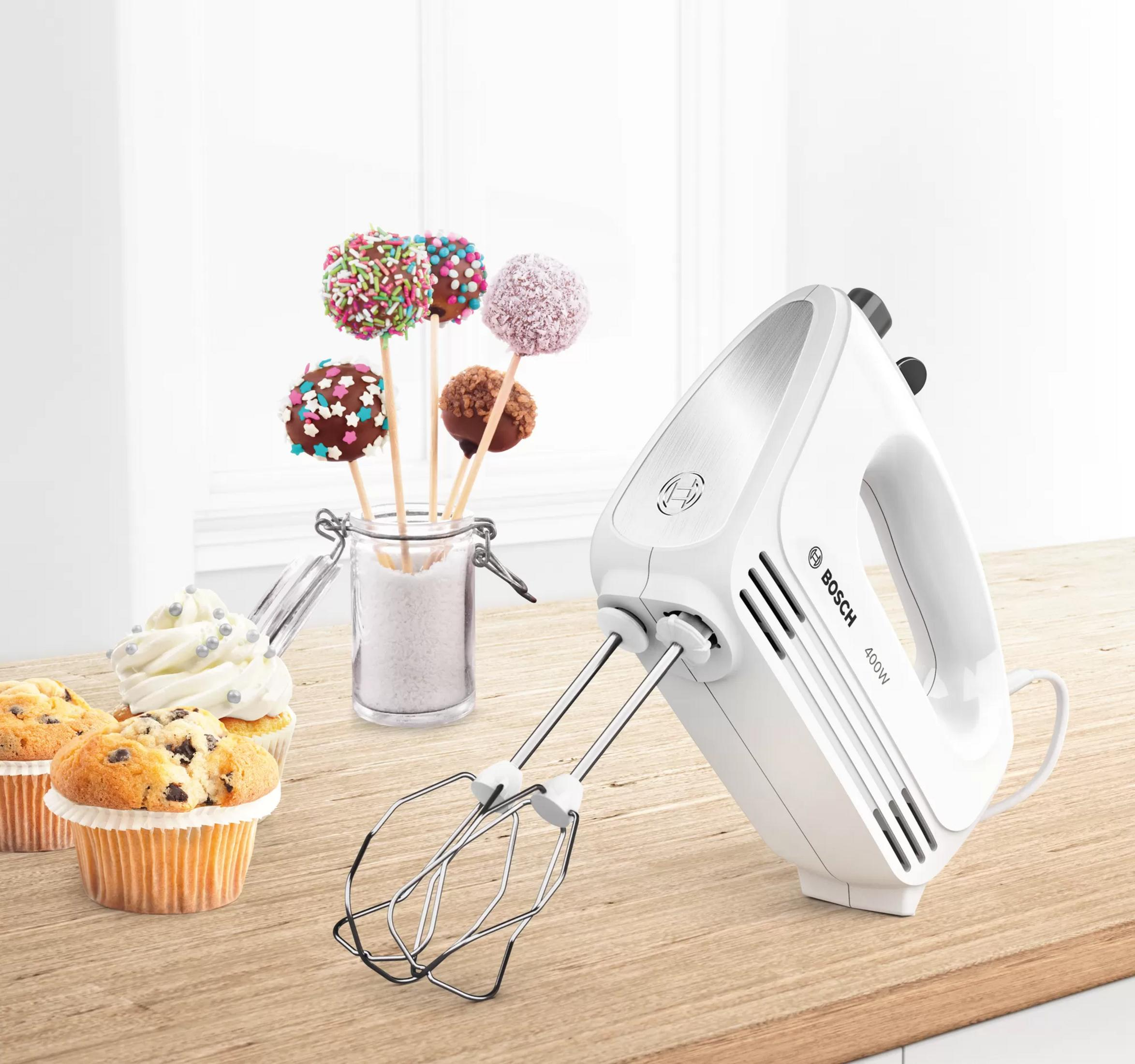 Ein weißer Bosch Handmixer mit Schneebesen, daneben Cupcakes und Cake Pops.