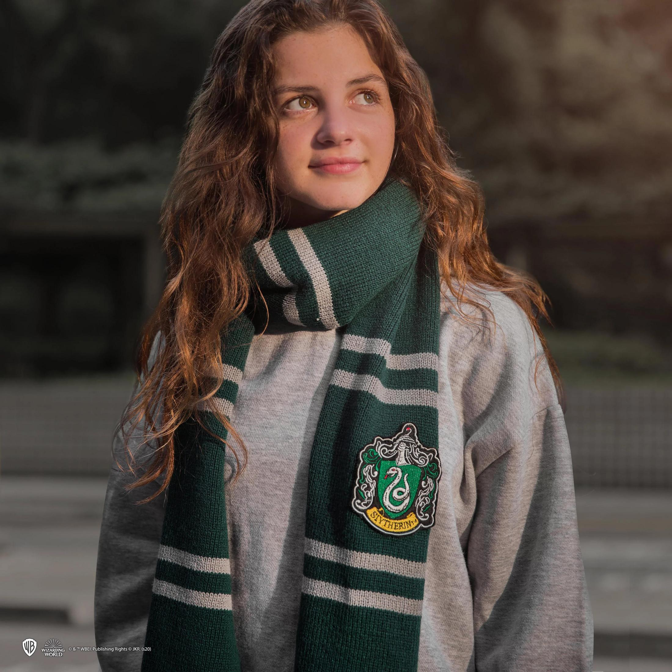 Ein Mädchen mit langen braunen Haaren trägt einen grauen Pullover und einen Schal mit Slytherin-Wappen.