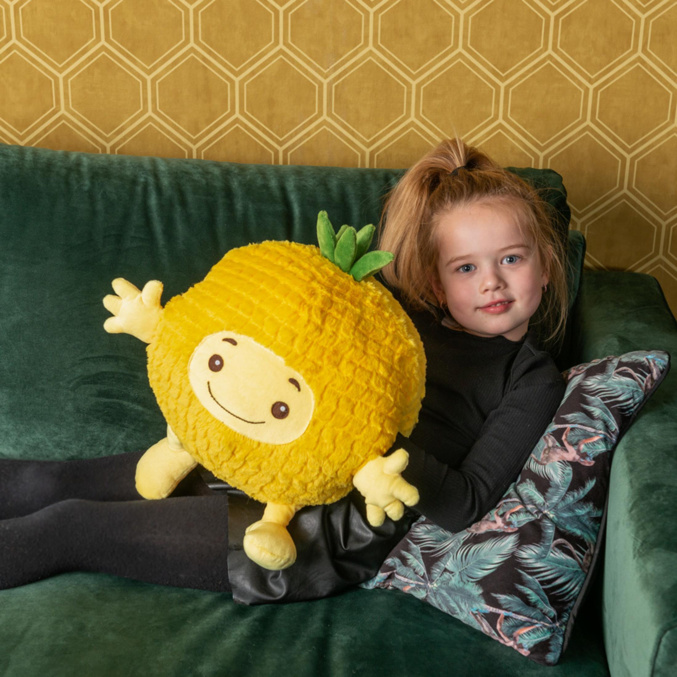 Ein kleines Mädchen sitzt mit einem gelben Ananas-Plüschtier auf einem grünen Sofa.