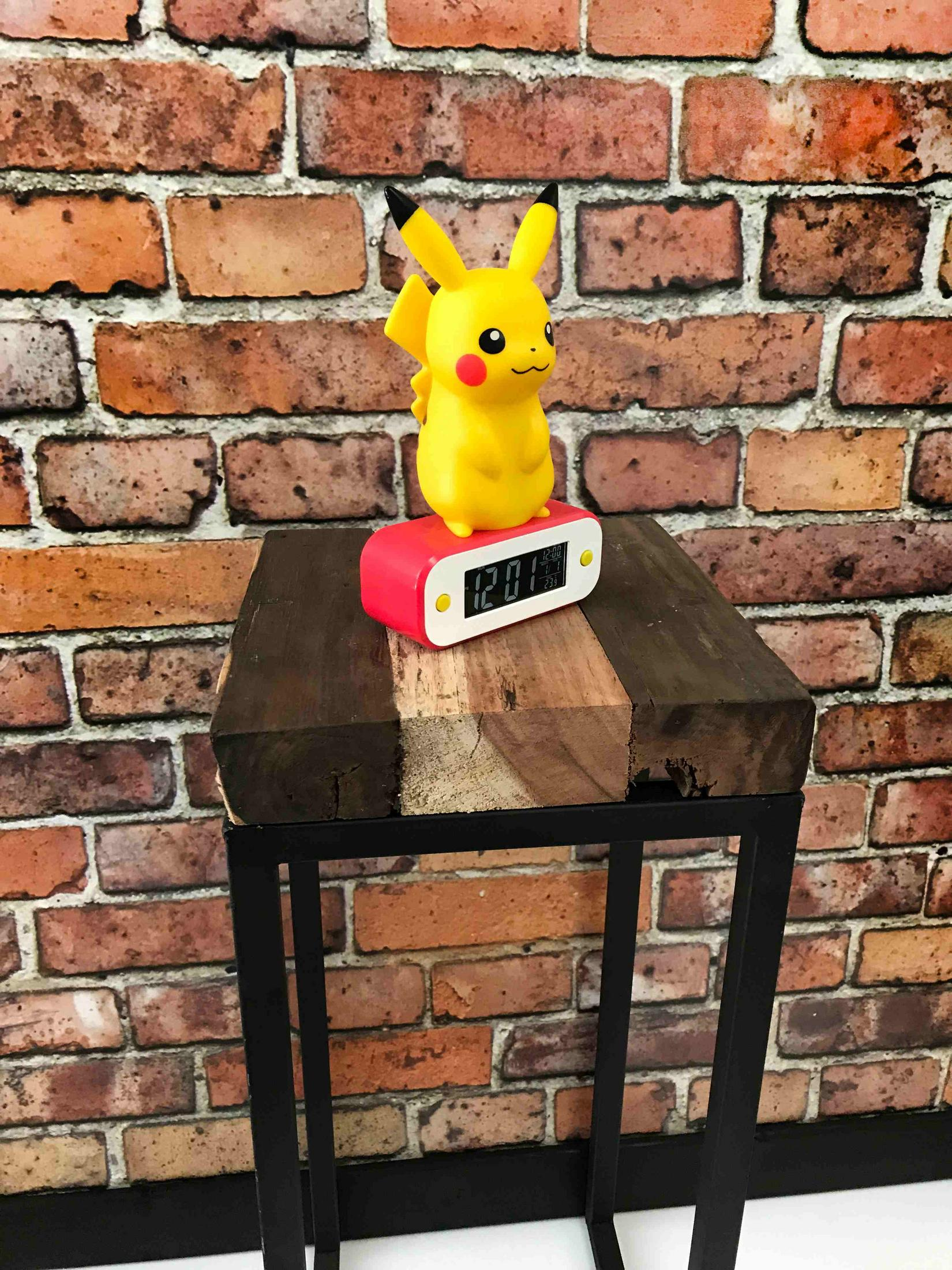 Figura gialla di Pikachu su sveglia rossa su tavolo di legno contro muro di mattoni.