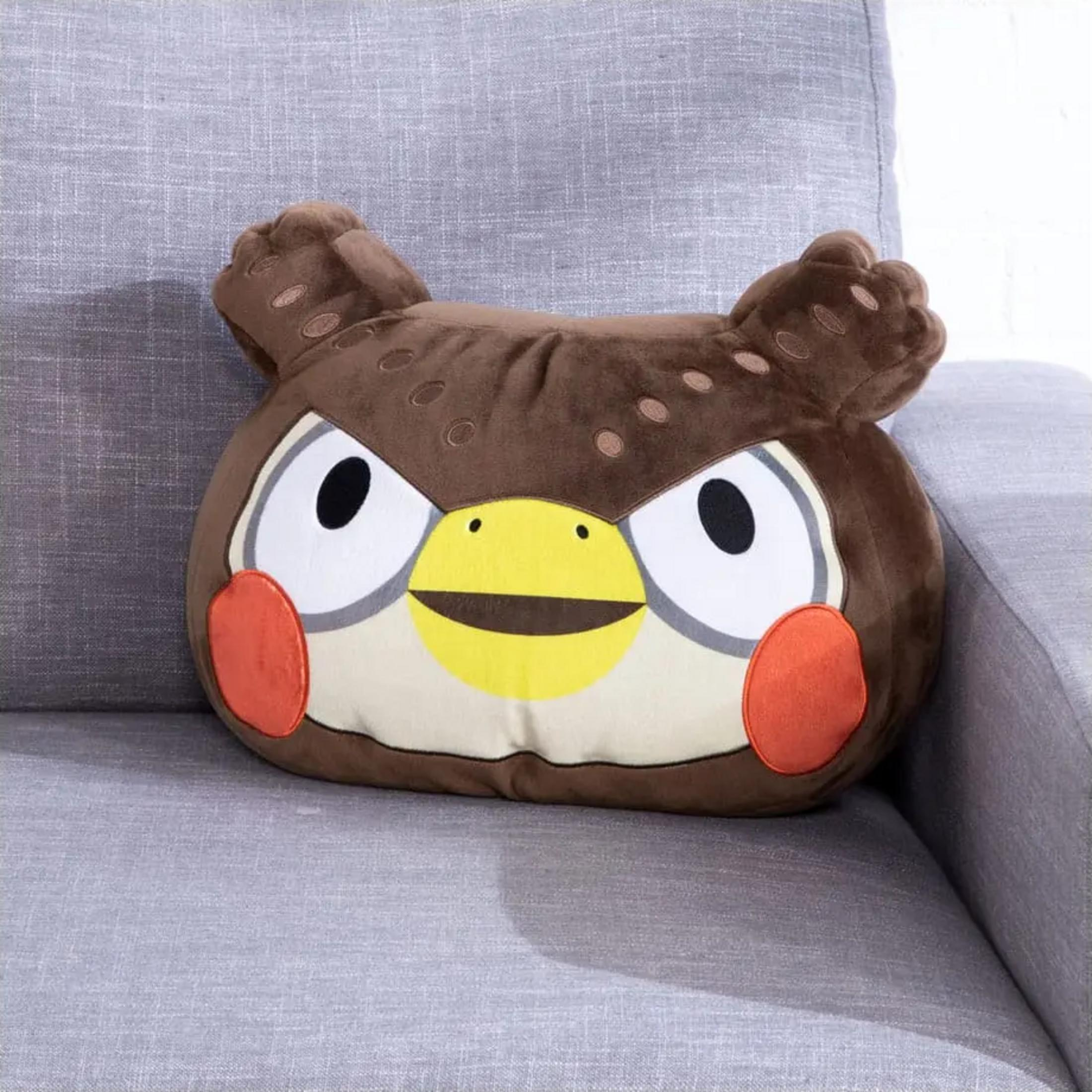 Ein braunes Eulen-Kissen mit großen Augen und roten Wangen liegt auf einem grauen Sofa.