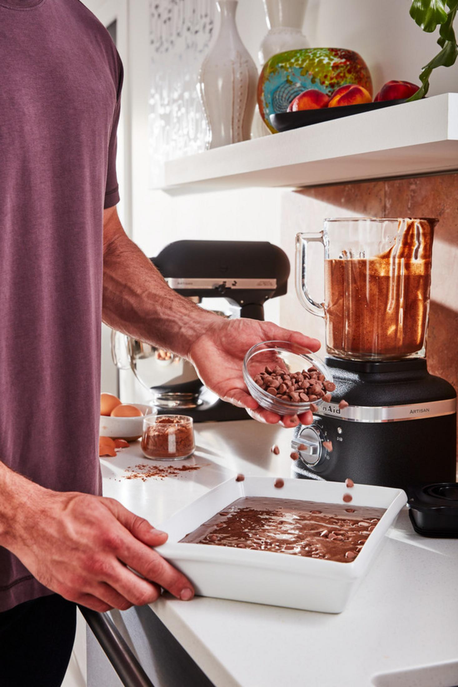 Person gibt Schokoladenstückchen zu Teig in weißer Schale. Mixer und Küchenmaschine im Hintergrund.