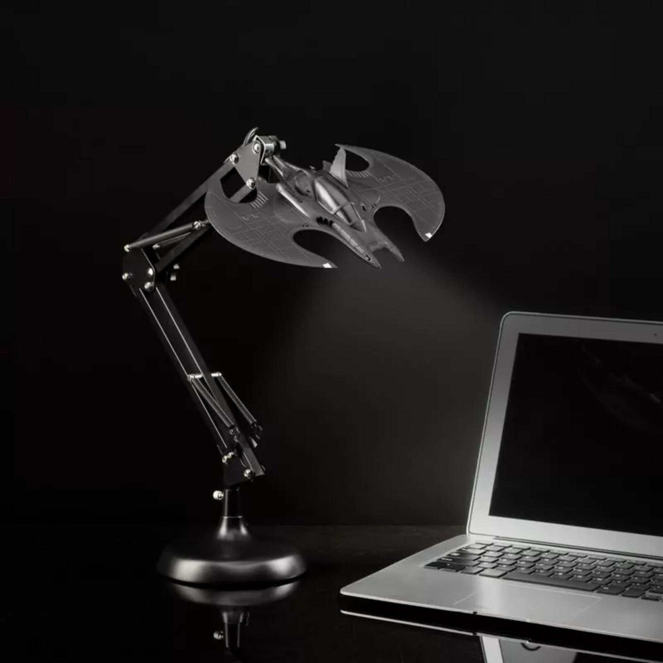 Ein schwarzer Schreibtischlampenarm mit einem Fledermaus-Schiff als Lampe leuchtet auf einen Laptop.