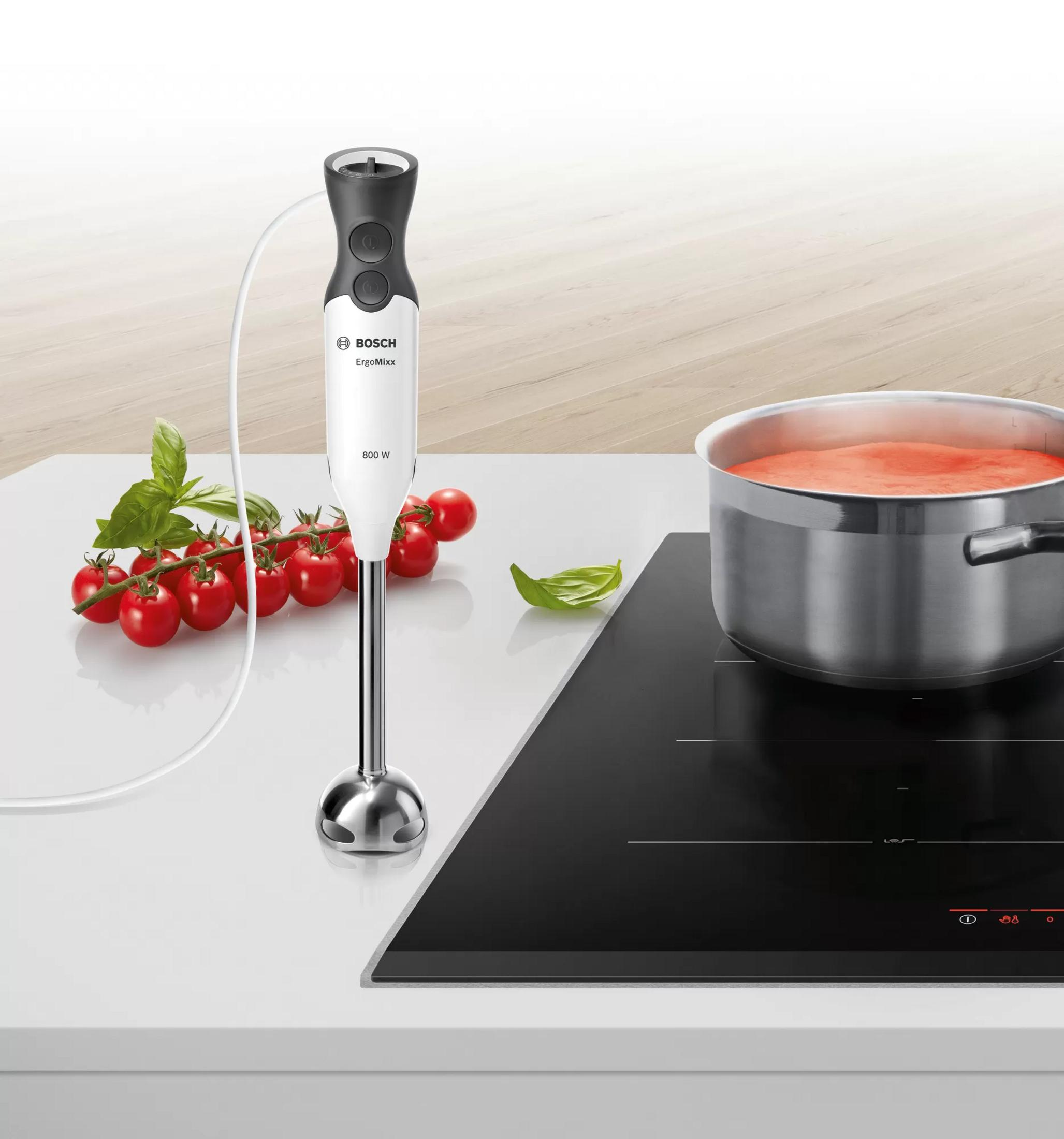 Biały blender ręczny Bosch z kablem, obok pomidorów i garnka z czerwoną cieczą.