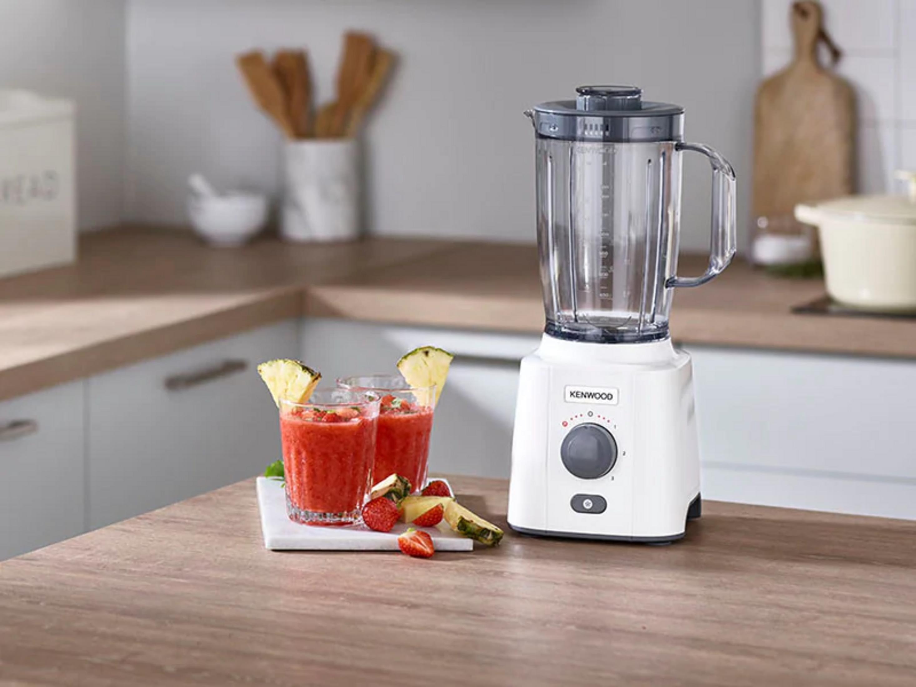 Beyaz bir Kenwood blender, ananas ve çilek ile hazırlanmış iki bardak çilekli smoothie yanında duruyor.