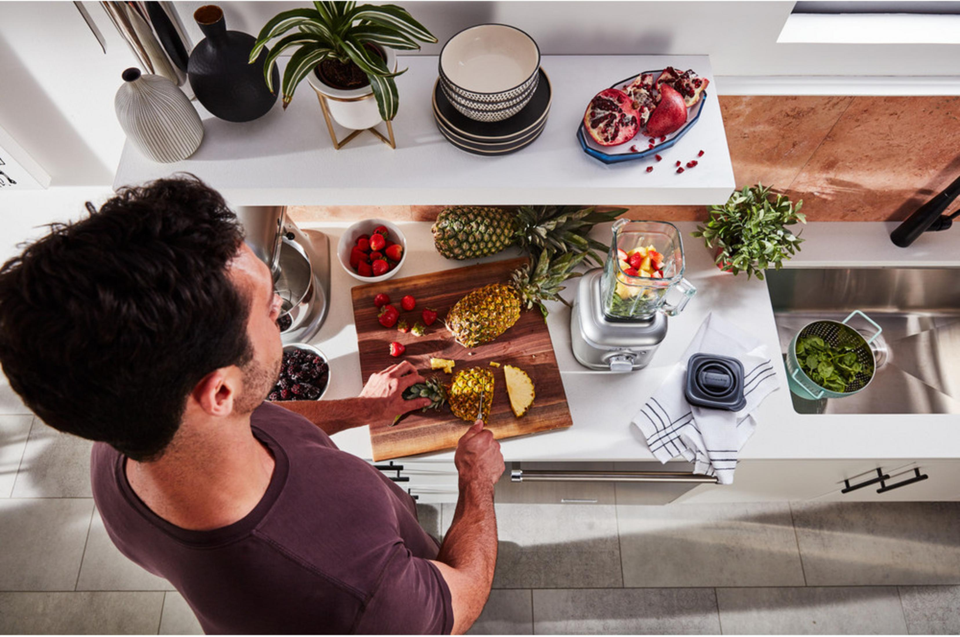 Mężczyzna kroi ananasa na desce, obok truskawek i blendera.