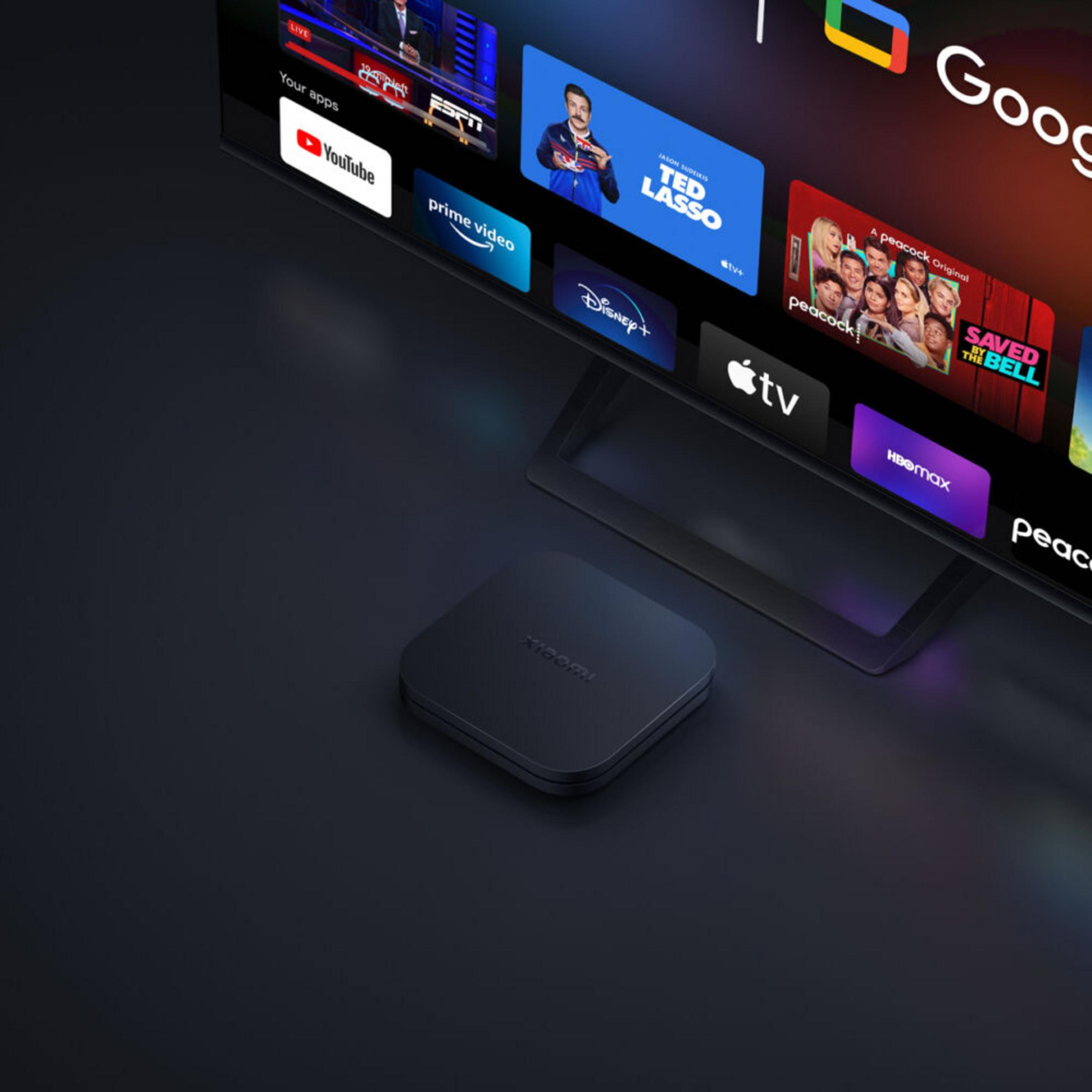 Una caja Xiaomi TV negra junto a un televisor que muestra iconos de aplicaciones.
