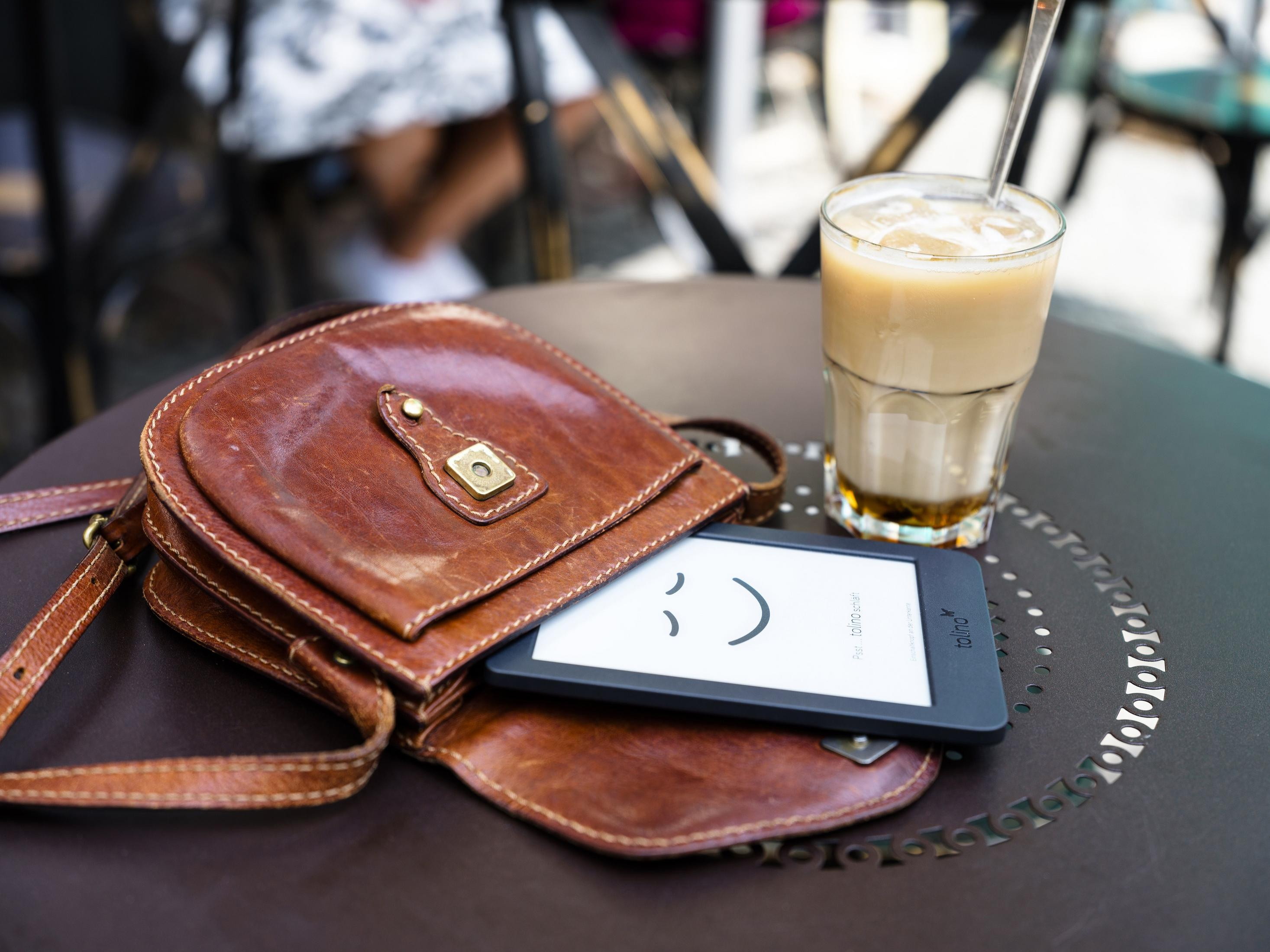 Eine braune Ledertasche mit einem E-Reader und einem Eiskaffee auf einem Tisch.