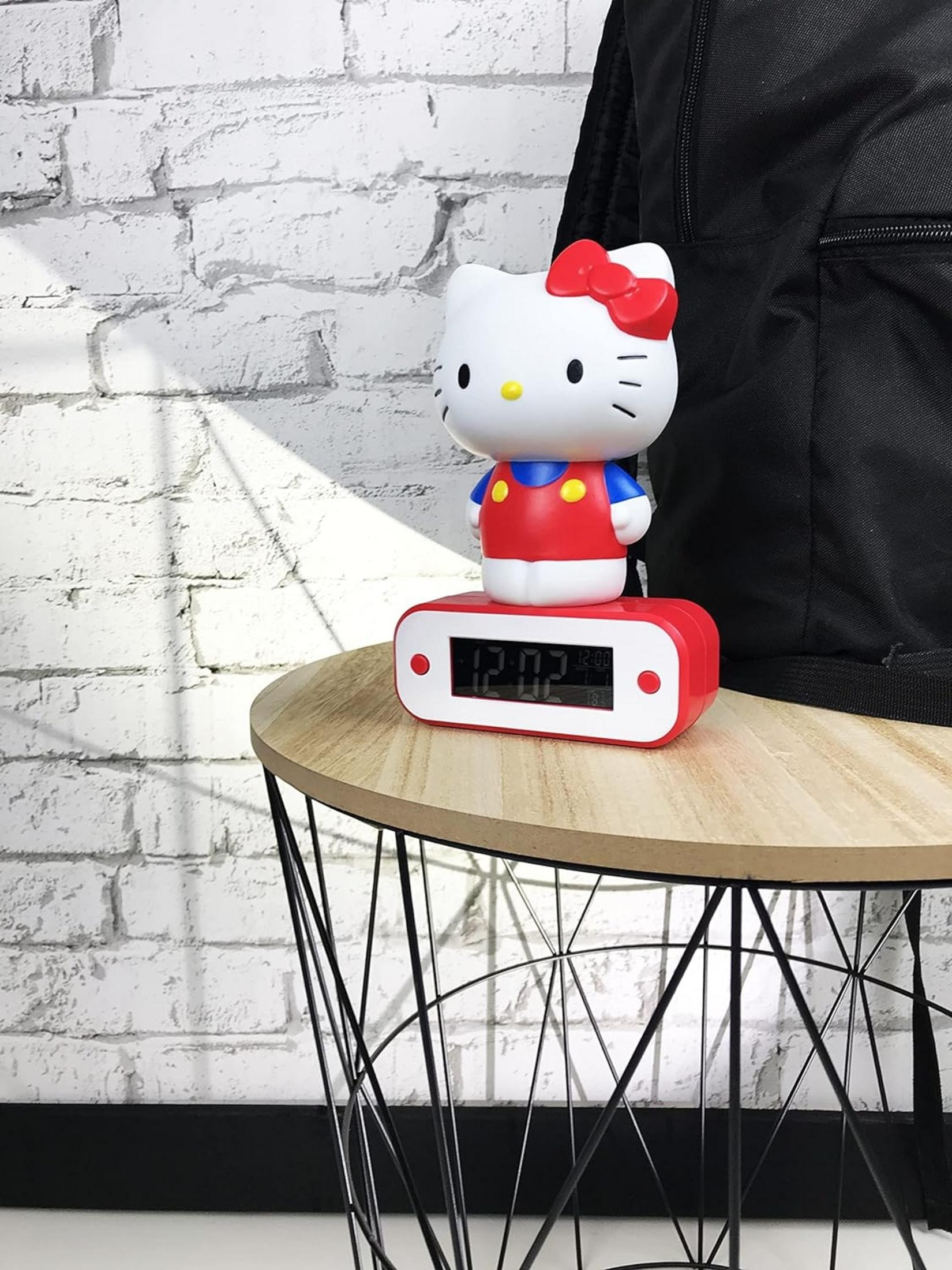 Budzik Hello Kitty stoi na drewnianym stole na tle białej ceglanej ściany.
