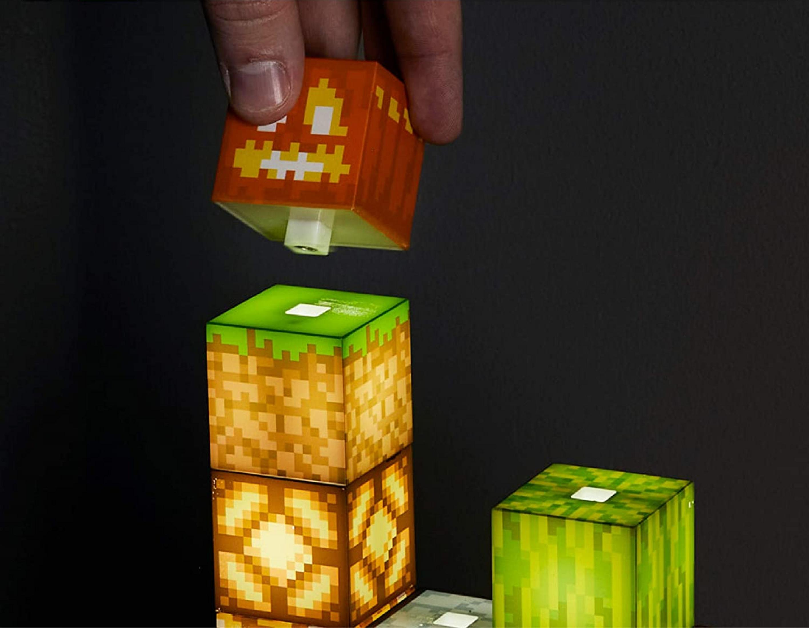 Ein Finger hält einen orangen Würfel mit gelbem Gesicht über leuchtende Minecraft-Blöcke.