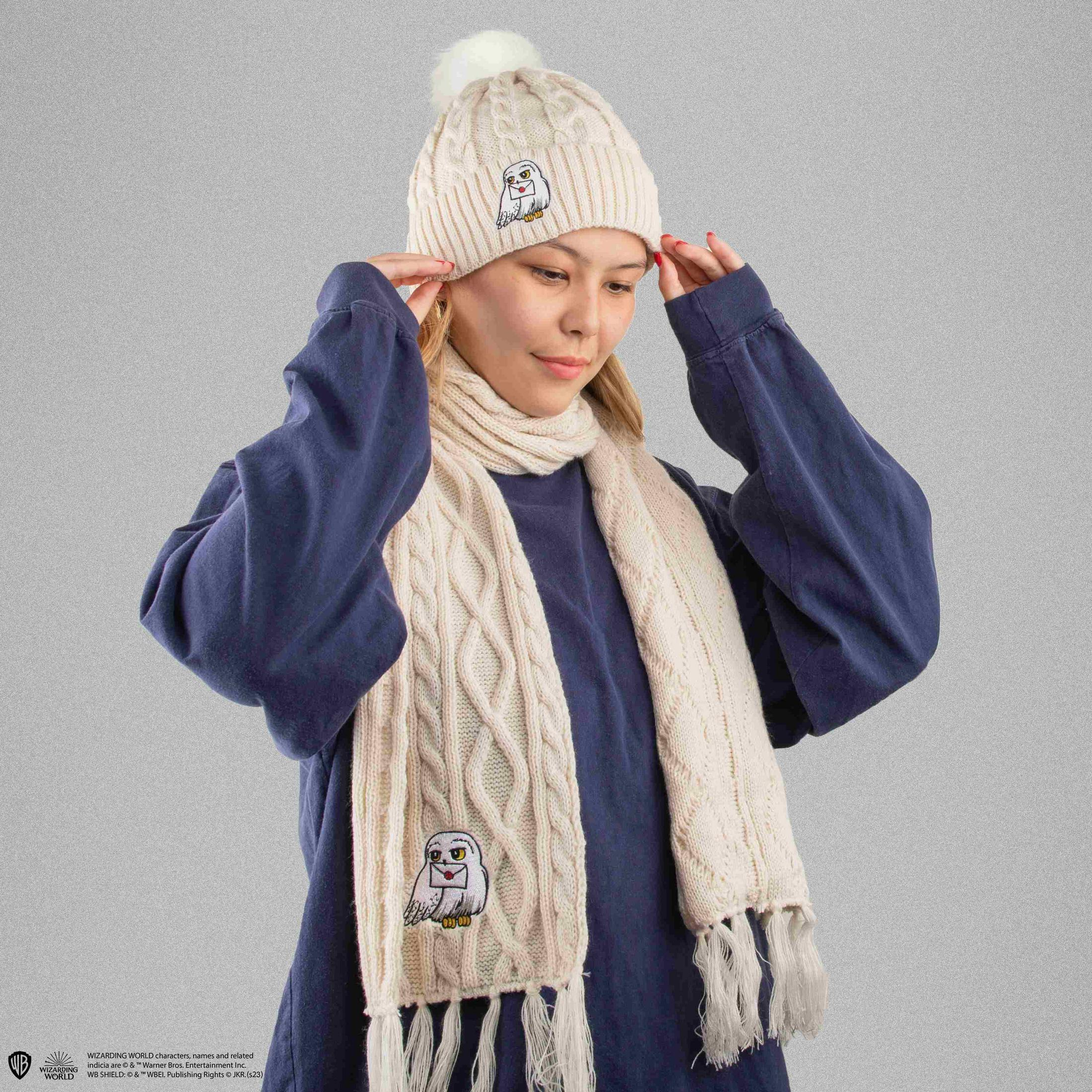 Eine Frau trägt eine weiße Strickmütze mit Bommel und einen passenden Schal mit Eulenmotiv.