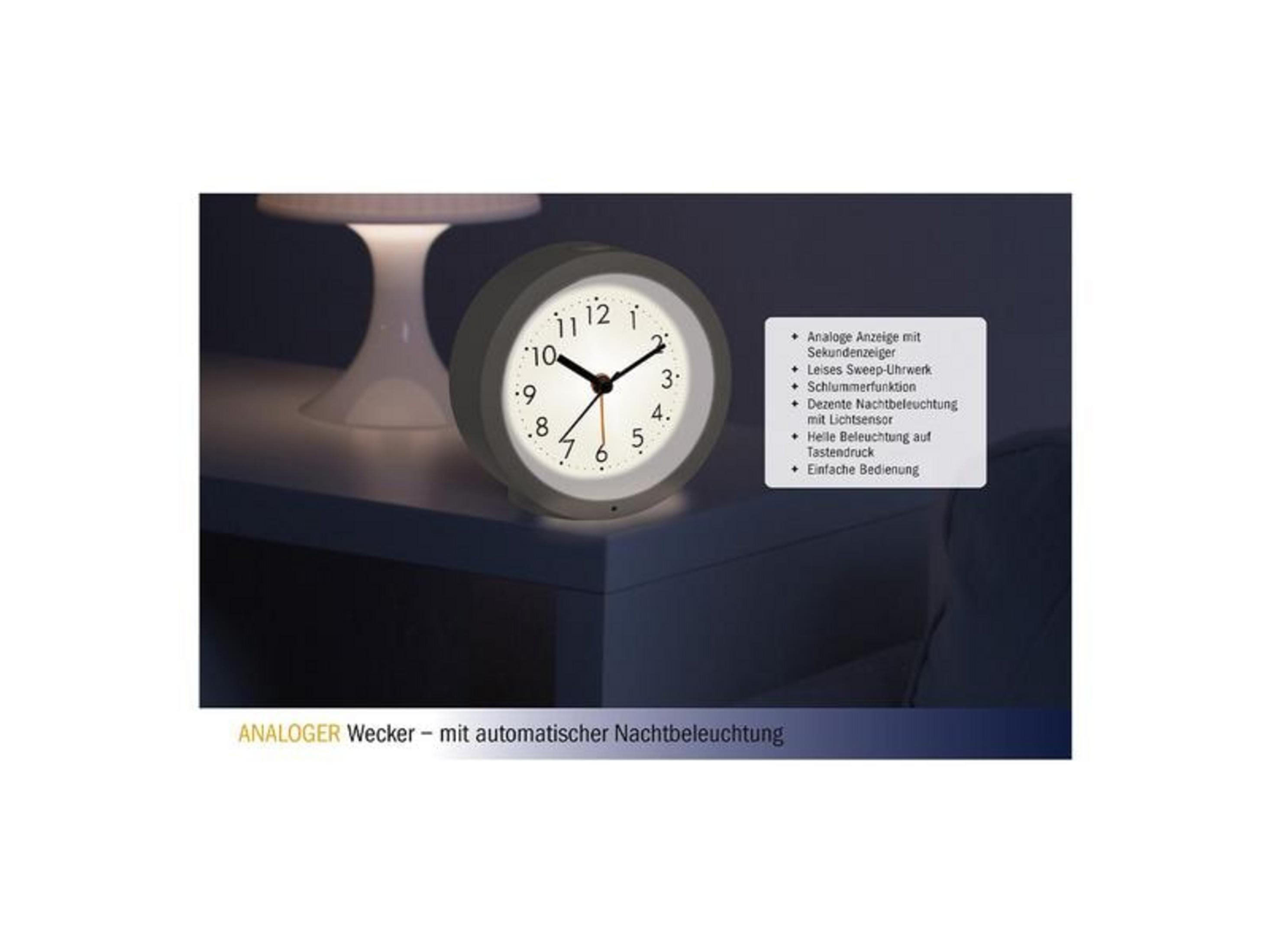 Un réveil gris avec un cadran analogique est posé sur une table à côté d'une lampe.