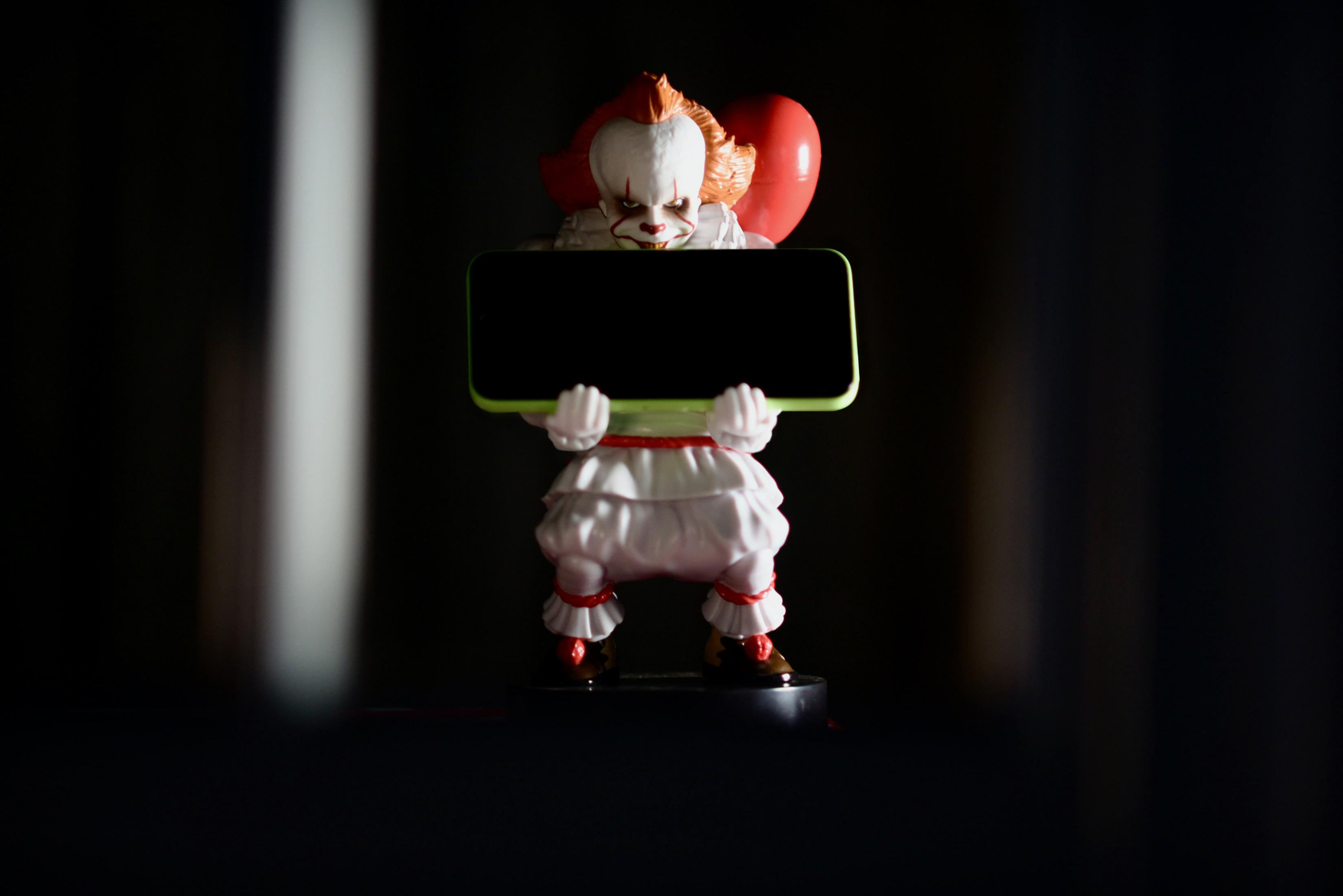 Eine gruselige Clown-Figur mit rotem Haar und rotem Ballon hält ein schwarzes Rechteck.