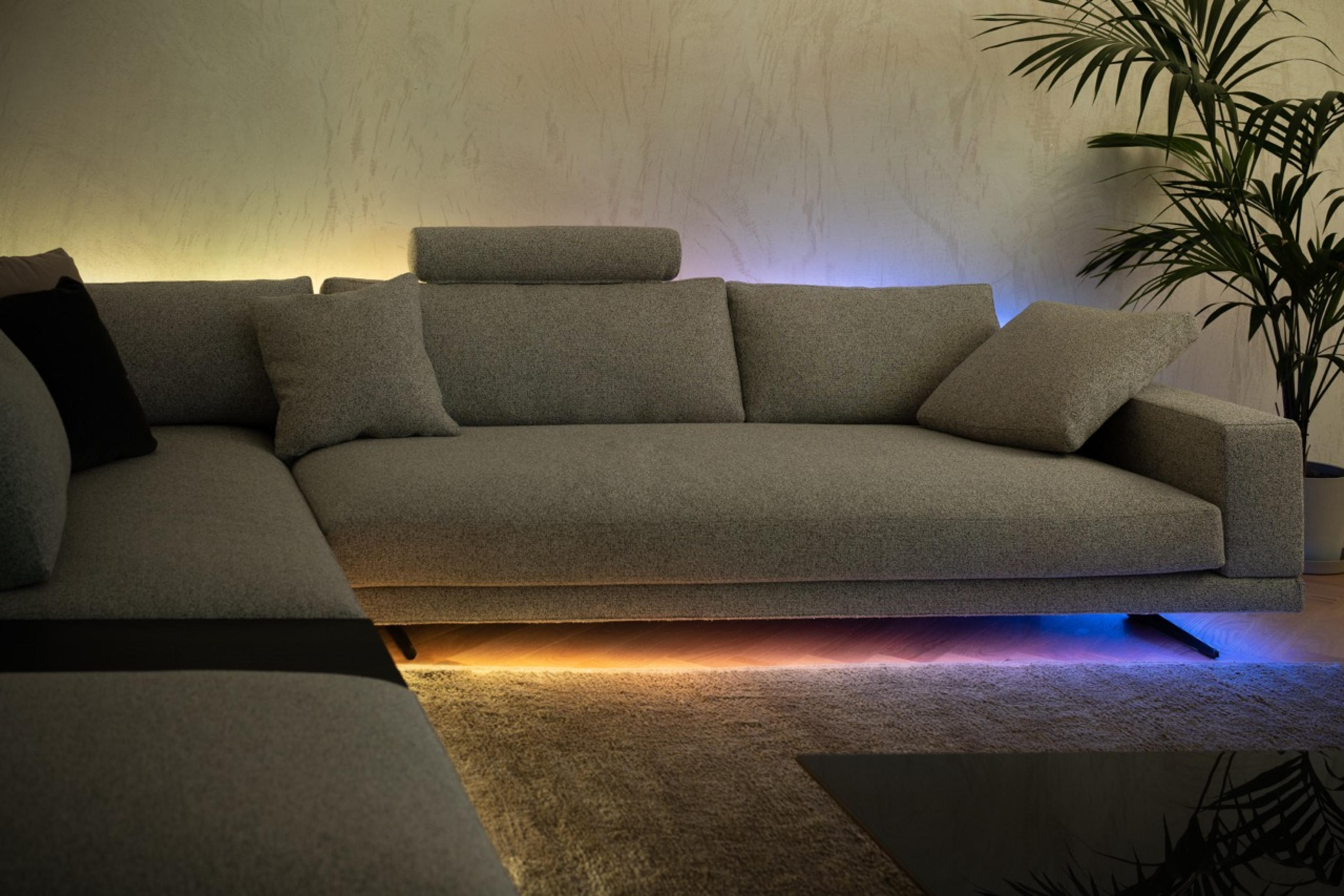 Szara sofa narożna z kolorowymi światłami LED pod spodem i rośliną.