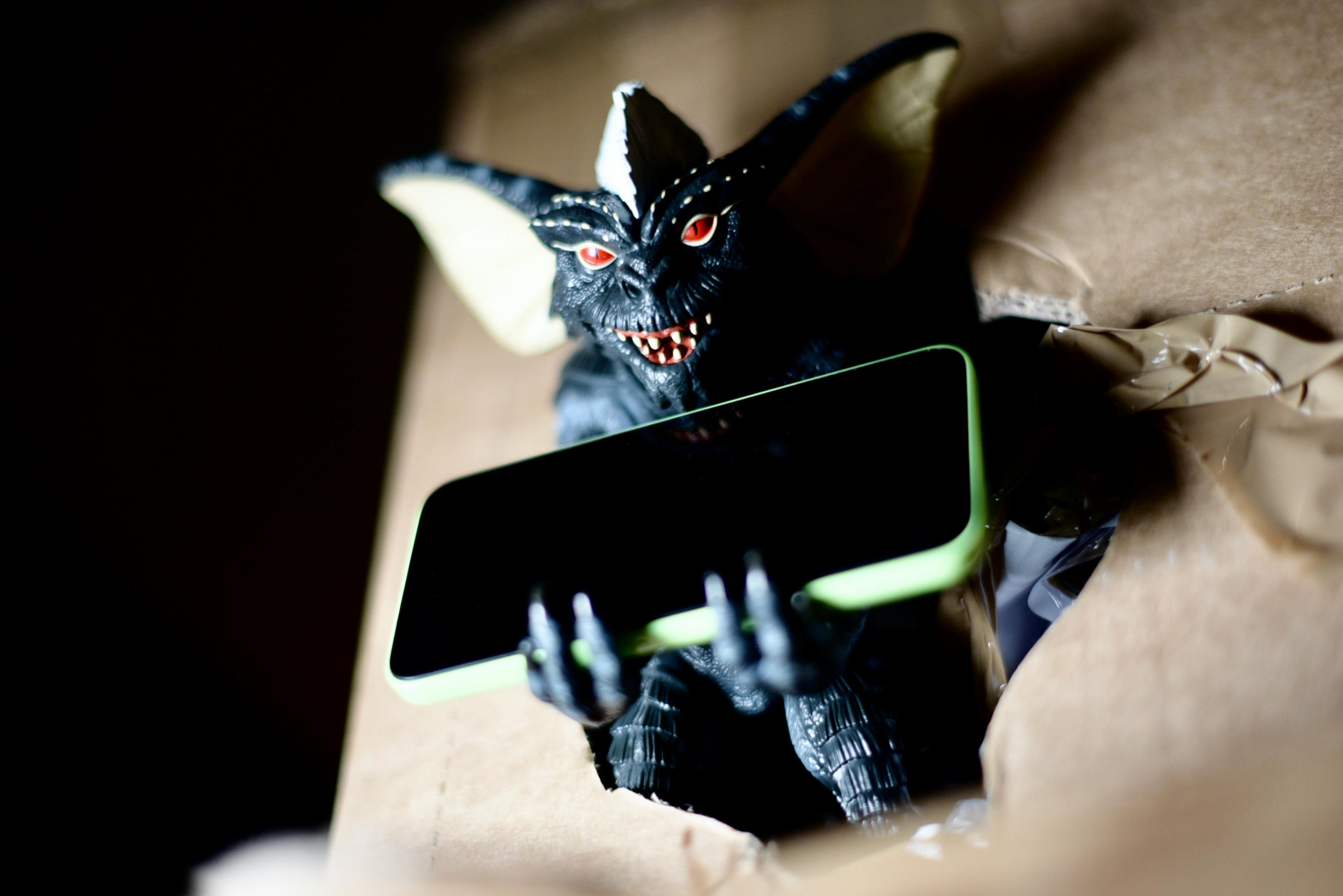 Un giocattolo Gremlin scuro tiene uno smartphone verde chiaro in una scatola di cartone.