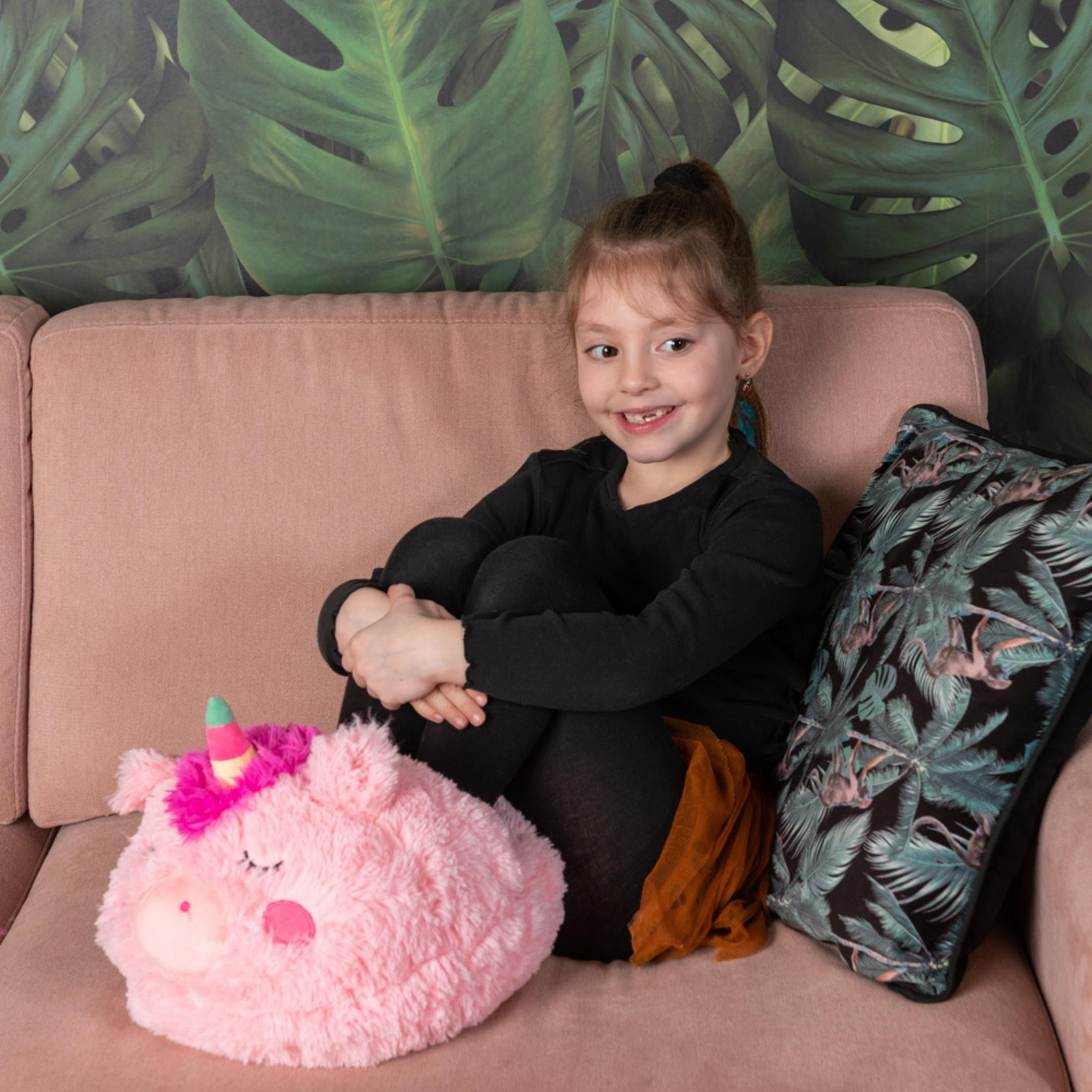 Ein kleines Mädchen sitzt auf einem Sofa neben einem Einhornkissen. Grüne Monstera-Tapete.