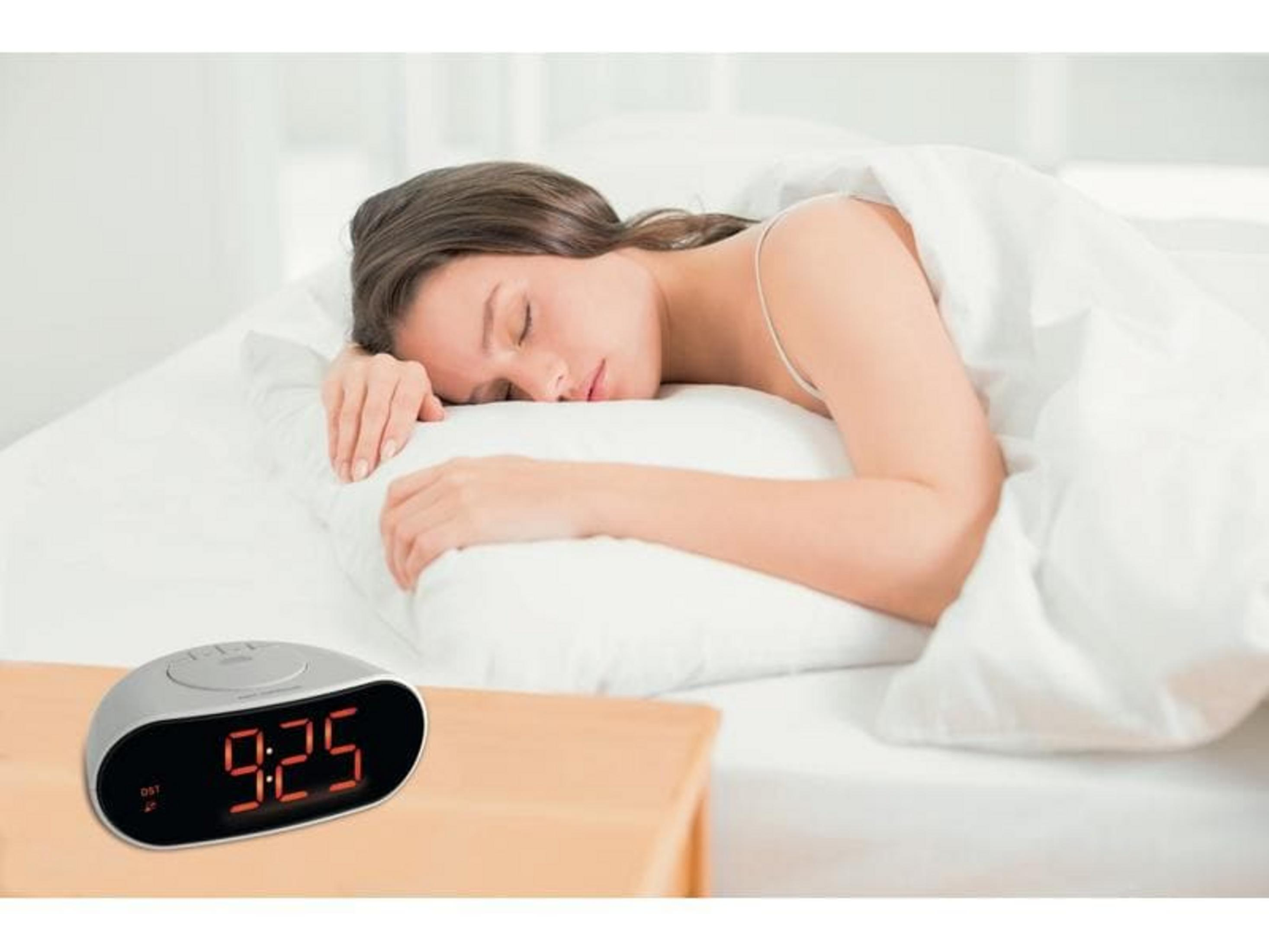 Eine Frau schläft im Bett, ein Wecker zeigt 9:25 an.