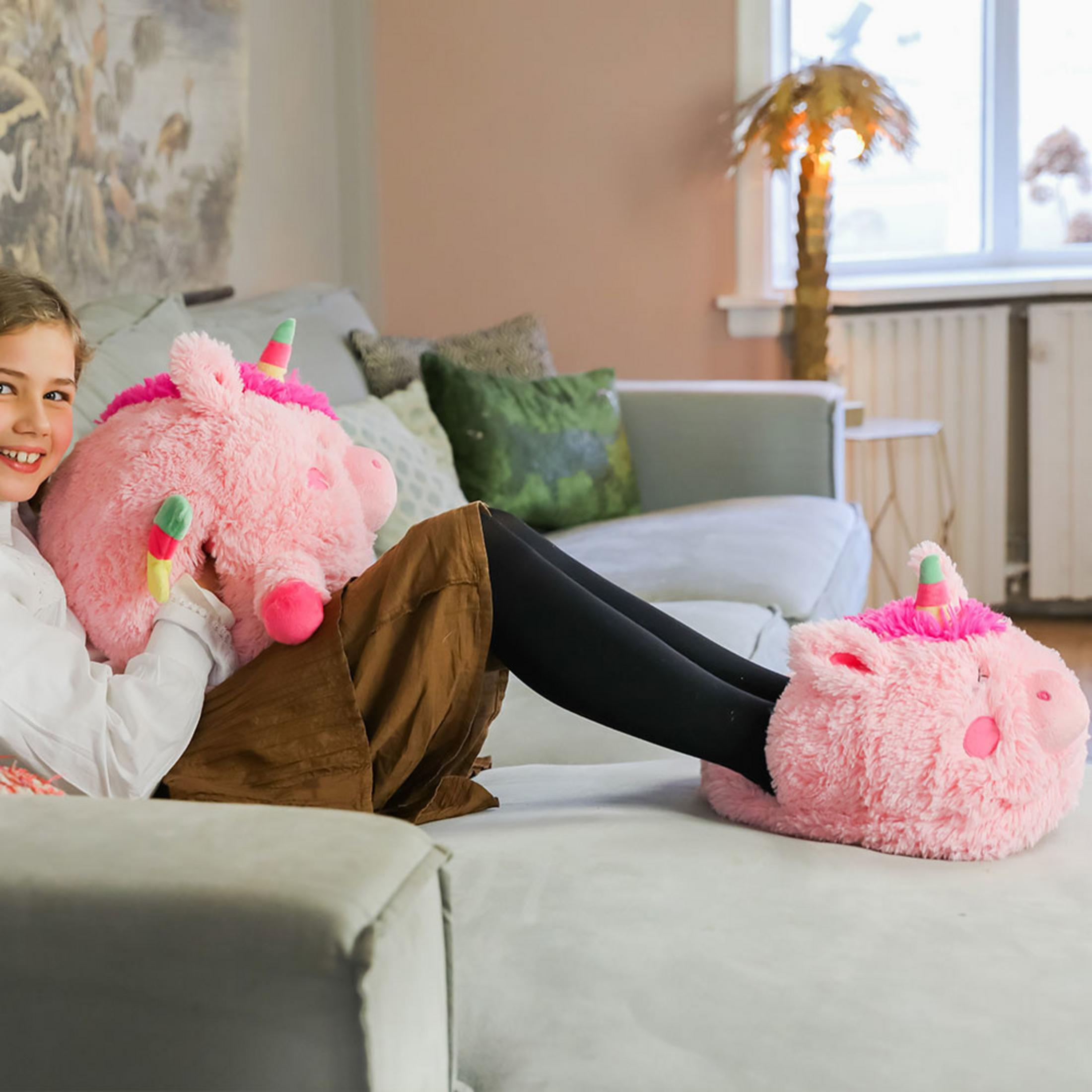 Ein Mädchen sitzt auf einem Sofa und hält ein rosa Einhorn-Kissen. Sie trägt Einhorn-Hausschuhe.