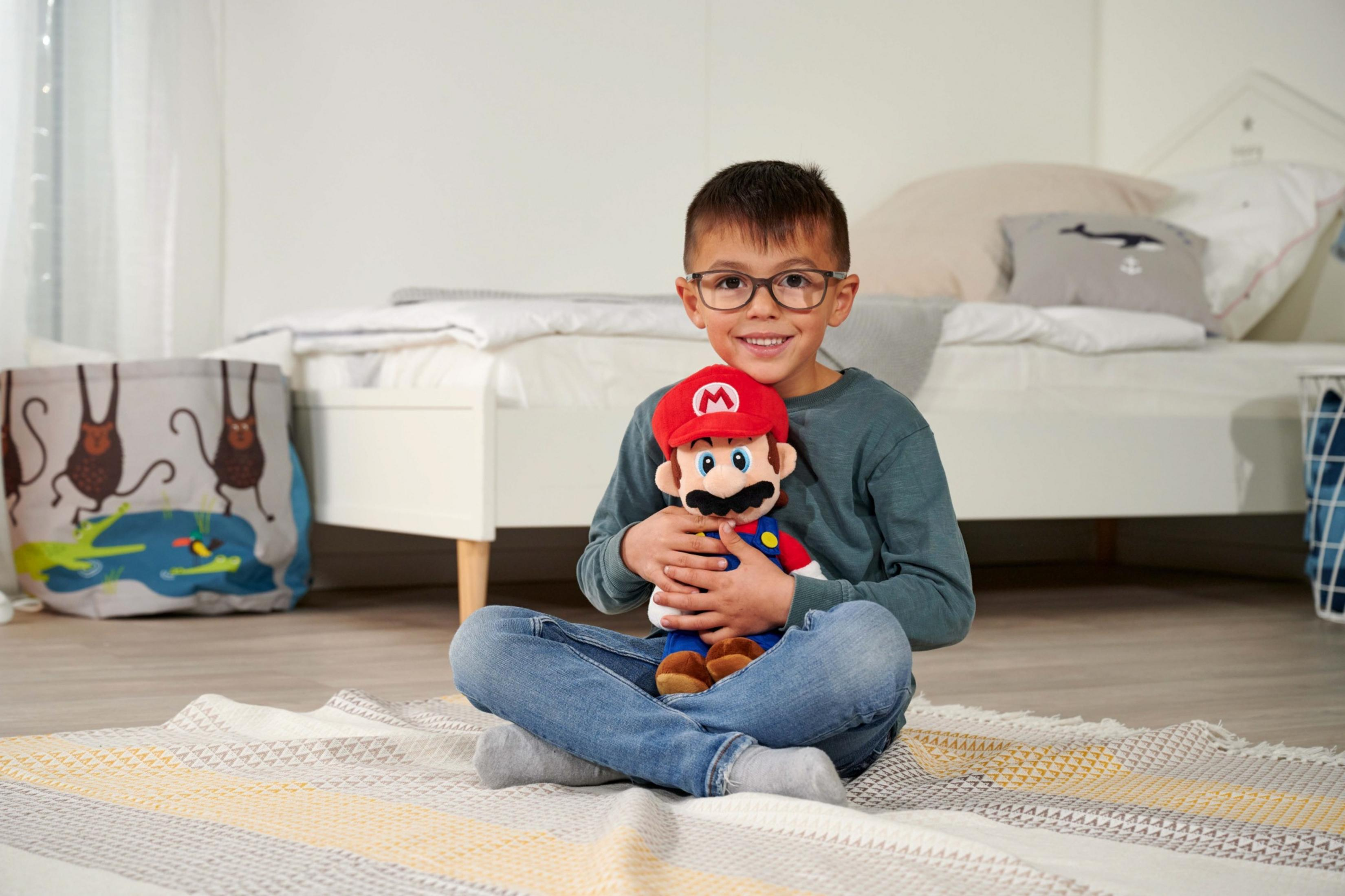 Ein Junge mit Brille sitzt auf einem Teppich und hält eine Mario-Puppe.
