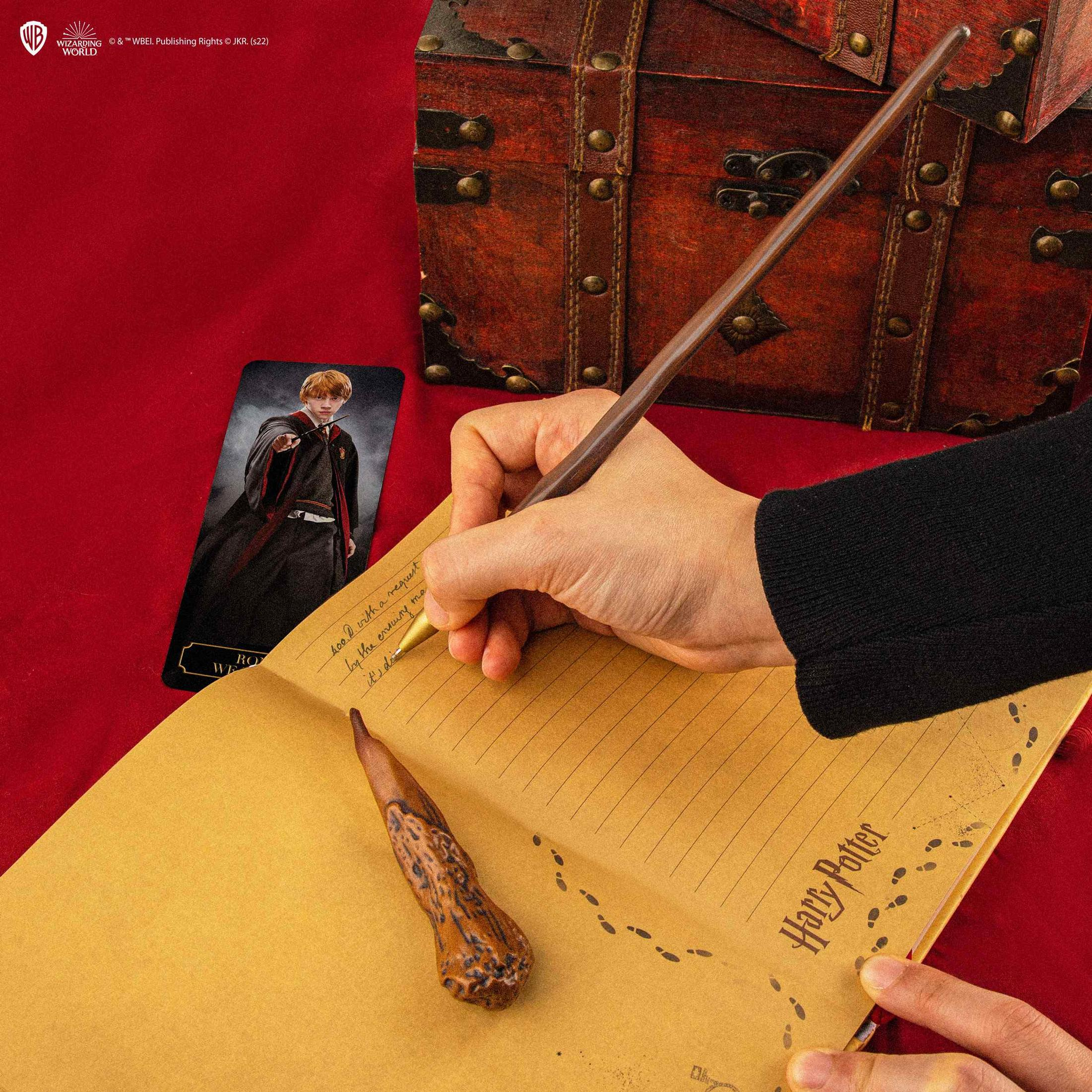Eine Hand schreibt in ein Notizbuch neben einer Zaubererkarte und einer Truhe.