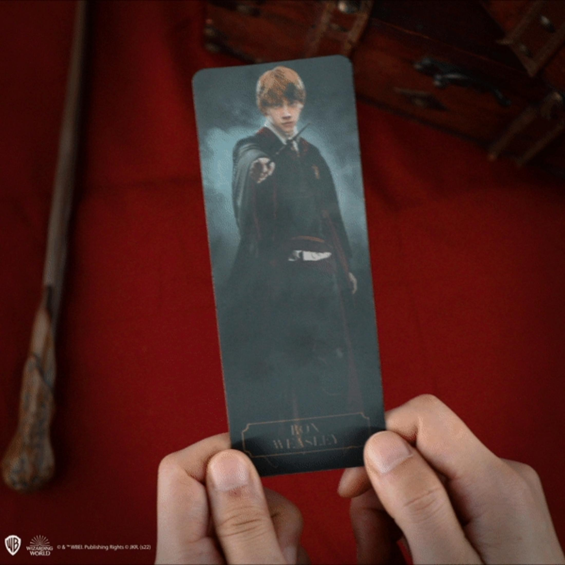 Ein Mann hält eine Lesezeichen mit Ron Weasley darauf. Ein Zauberstab liegt links.