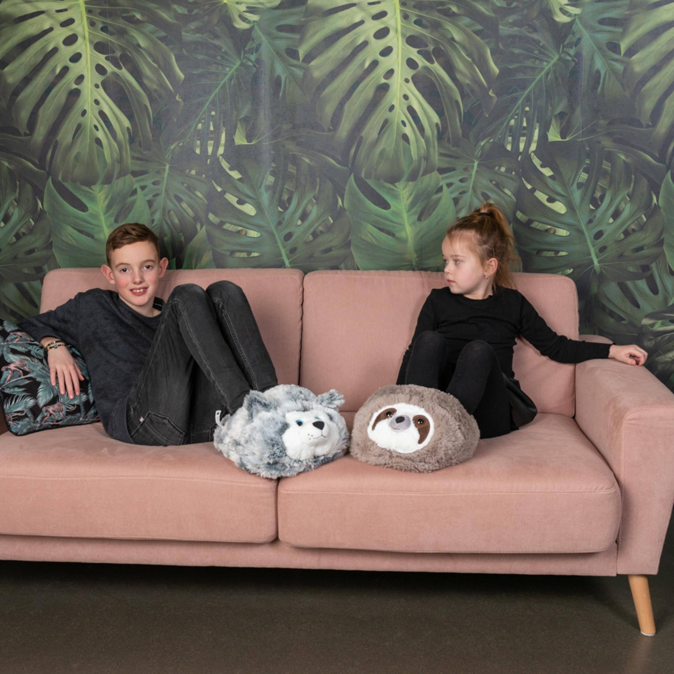 Kinder sitzen auf einem Sofa mit Monstera-Tapete. Sie tragen Tierpantoffeln.
