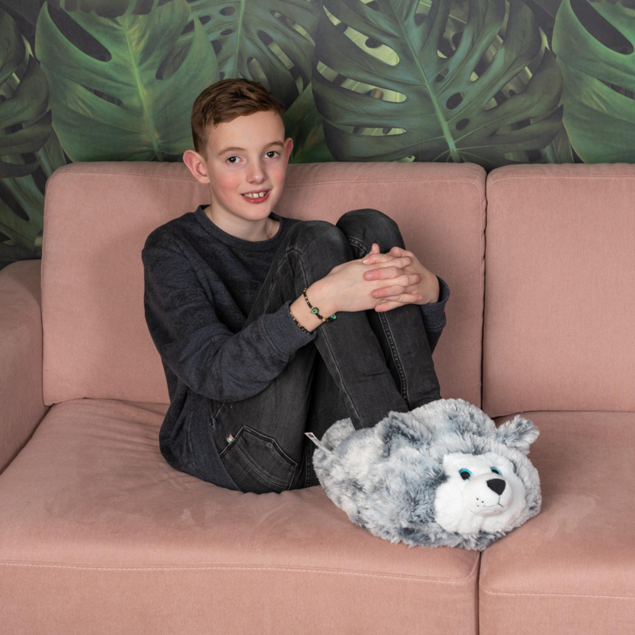 Junge sitzt auf rosa Sofa mit Plüschwolf-Hausschuhen vor Blatt-Tapete.